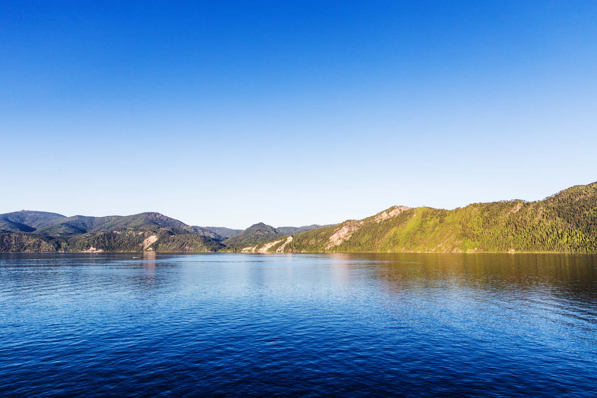 Lake 桌布 壁紙 — 寧靜的湖面倒映著清澈的藍天，遠處森林覆蓋的山脈在明亮的日光下聳立。