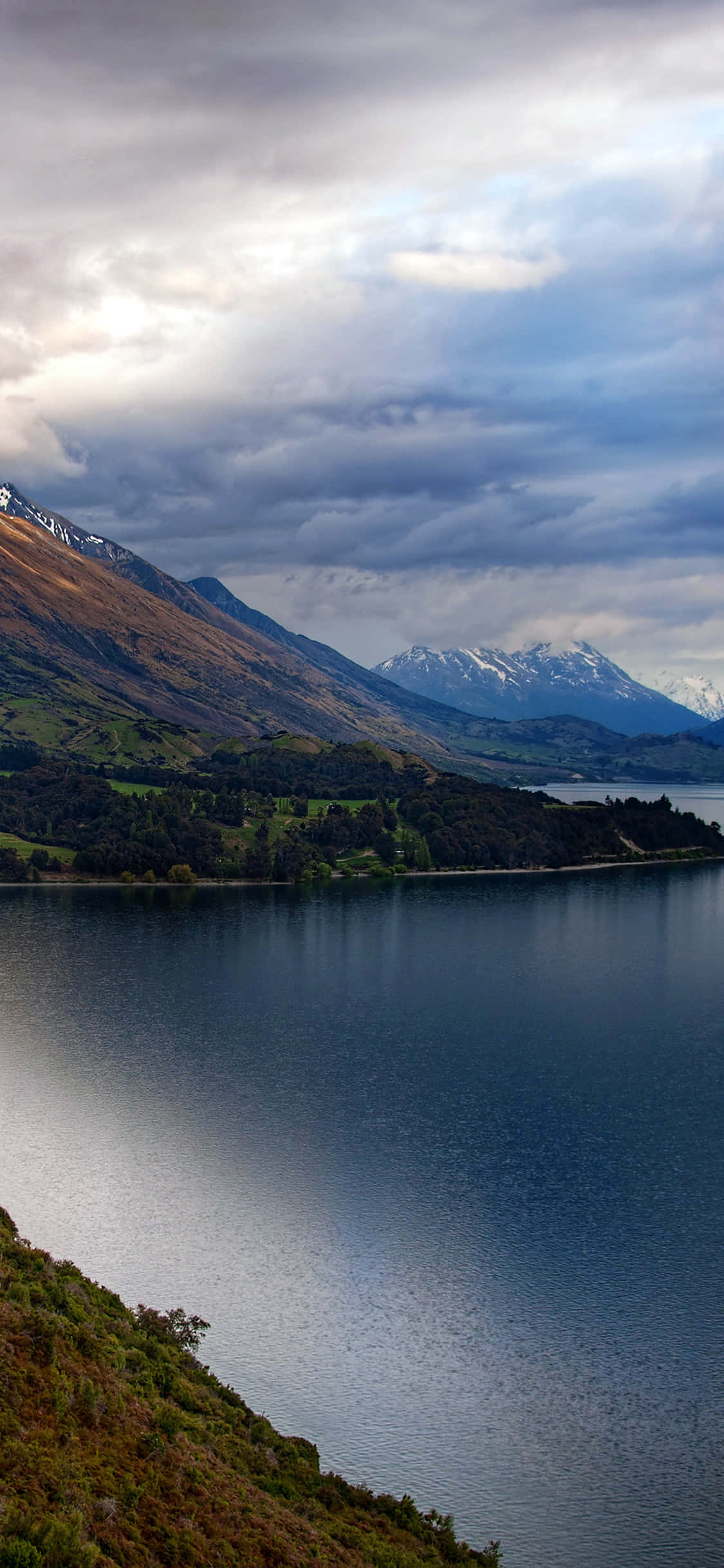 Beautiful Mountain Lake 桌布 壁紙 — 寧靜的湖面映照著戲劇性的多雲天空，白雪覆蓋的山脈在起伏的綠色山丘後方聳立。