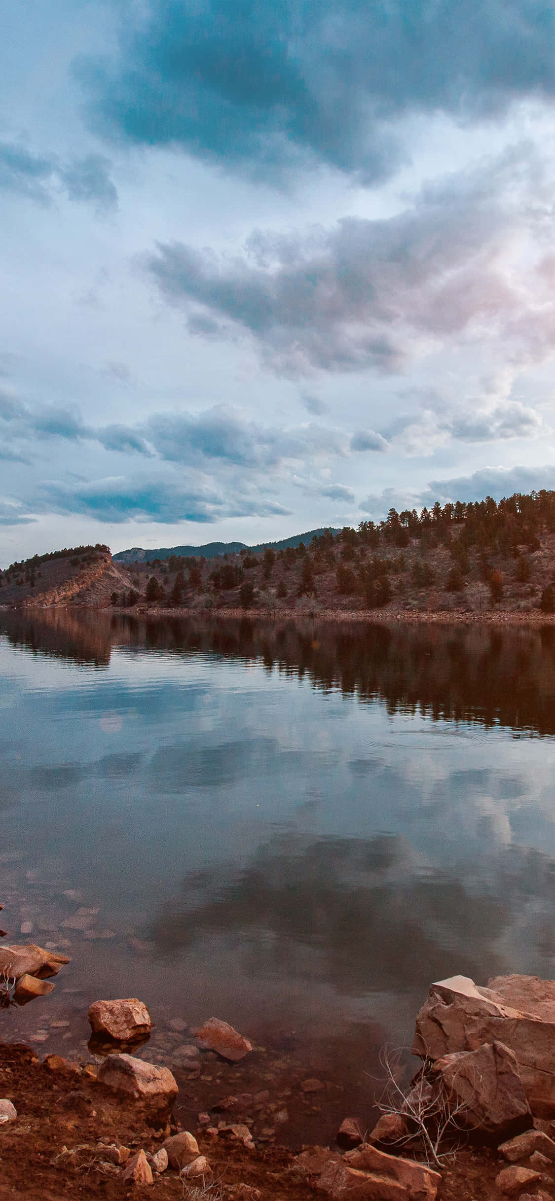 Lake 桌布 壁紙 — 寧靜的湖面映照著戲劇性的天空，雲朵飄過遠處樹木覆蓋的山丘和岩石海岸。