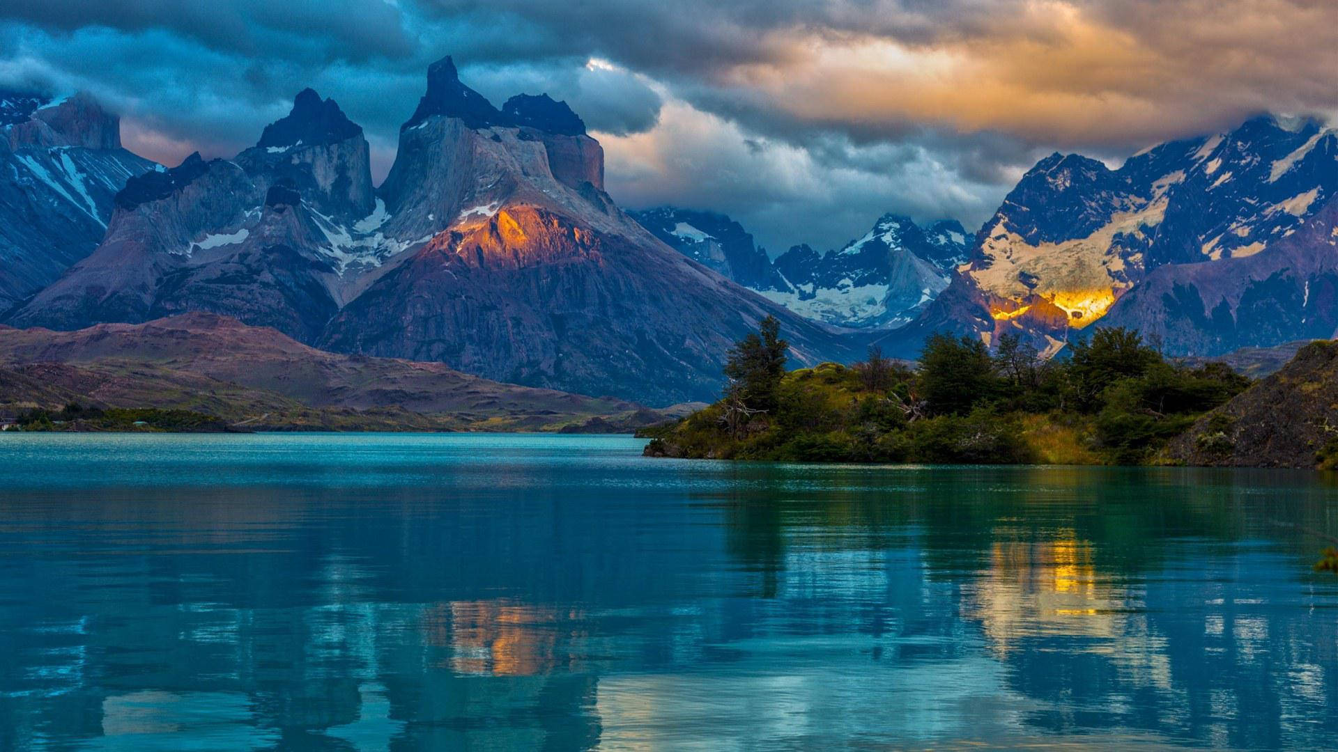 Argentina 桌布 壁紙 — 寧靜的湖面映照著白雪覆蓋的山峰，在戲劇性的雲層下閃耀著夕陽的色彩。
