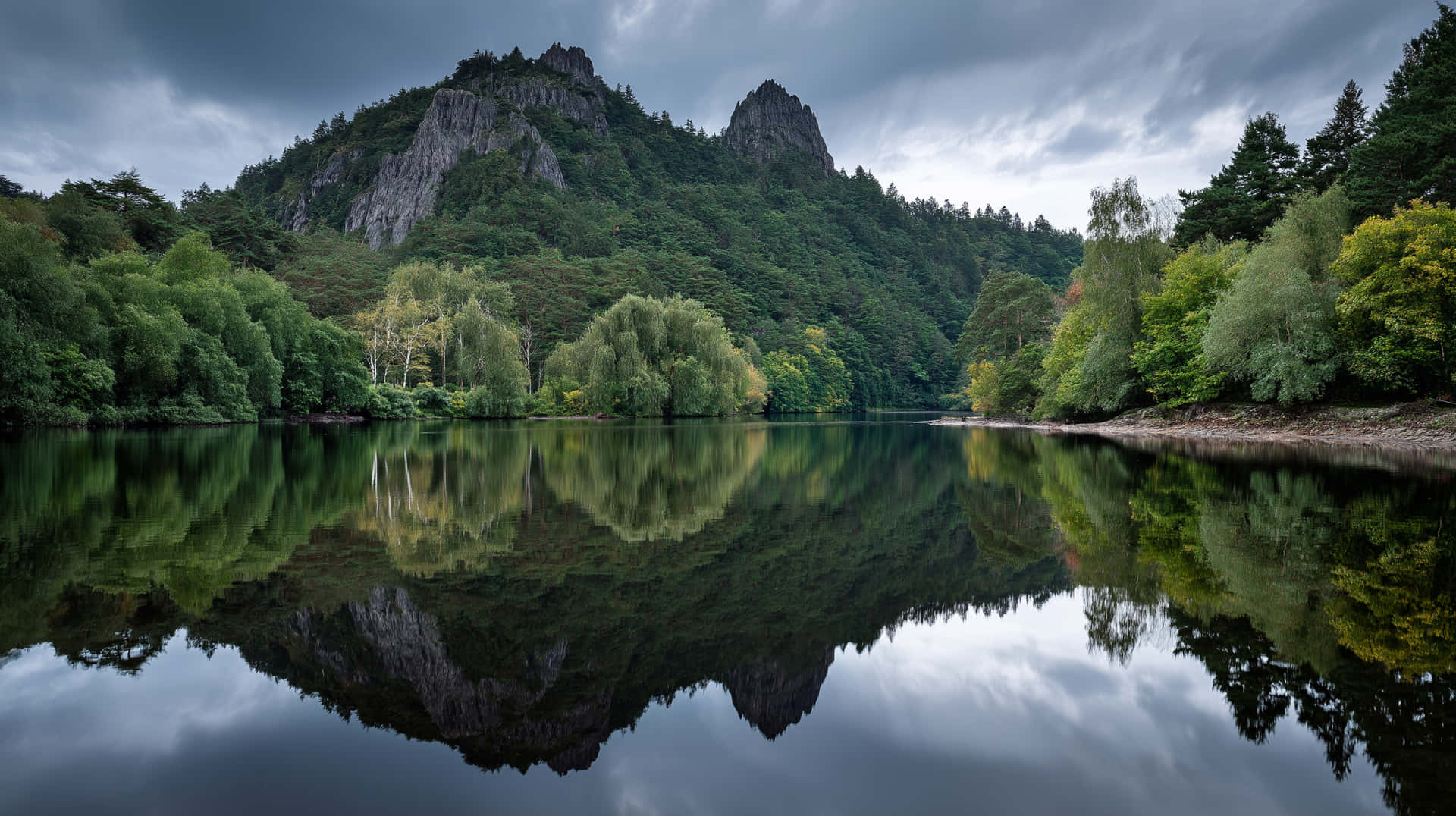 Mirrored Mountain Lake 壁紙 — 寧靜的湖面映照著陰沉灰天下崎嶇的森林覆蓋的山脈。