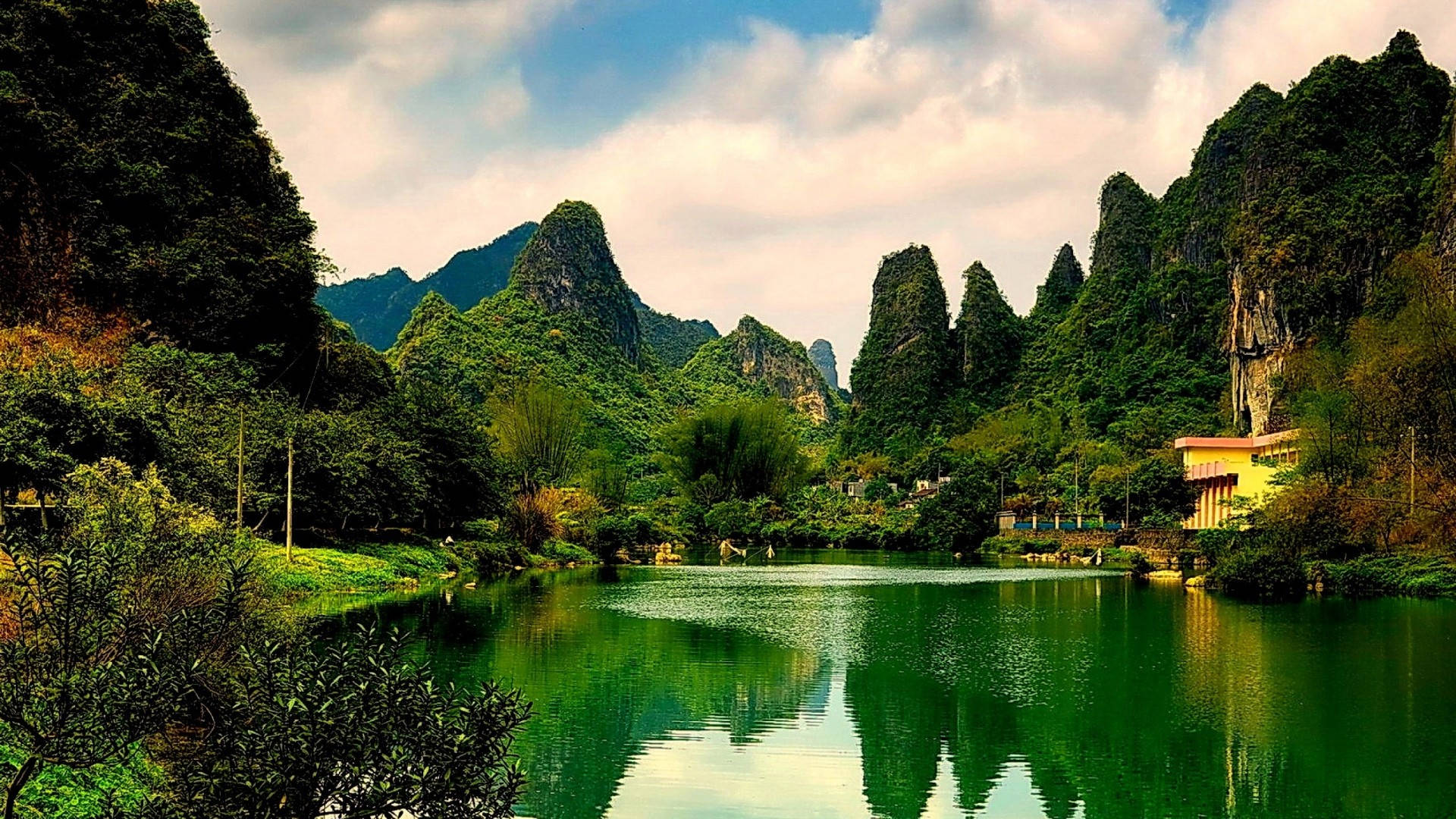 1920x1080 高畫質自然 桌布 壁紙 — 寧靜的湖面映照著鬱鬱蔥蔥的綠色喀斯特山脈，天空多雲，岸邊有一座小建築。