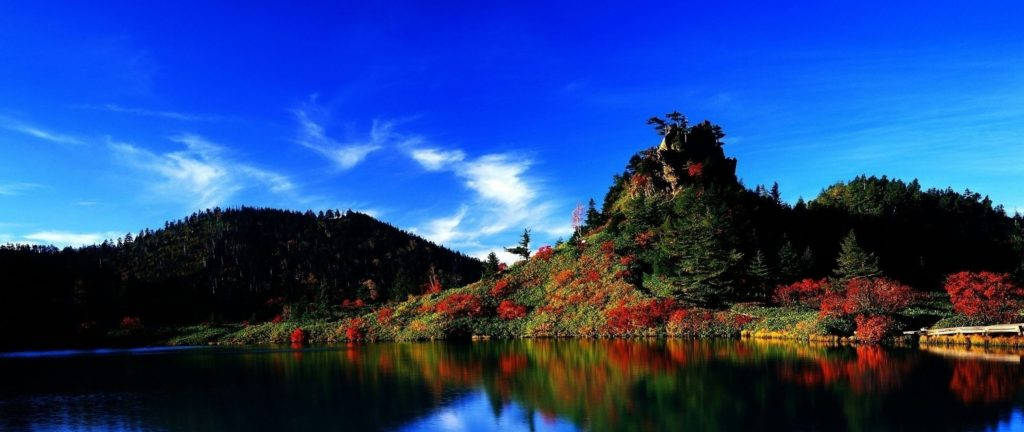 Beautiful Sky 桌布 壁紙 — 寧靜的湖面映照著鮮紅的秋葉和岩石山丘，天空深藍，雲朵輕柔。