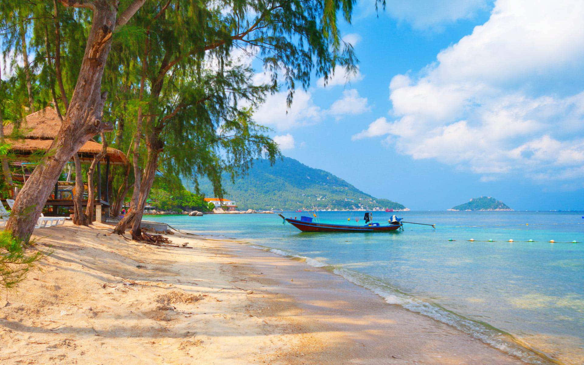 Thailand Beach 桌布 壁紙 — 寧靜的熱帶海灘，擁有碧綠海水、輕柔海浪、沙灘、茂密的綠樹，以及一艘在明亮藍天白雲下漂浮在岸邊的長尾船。