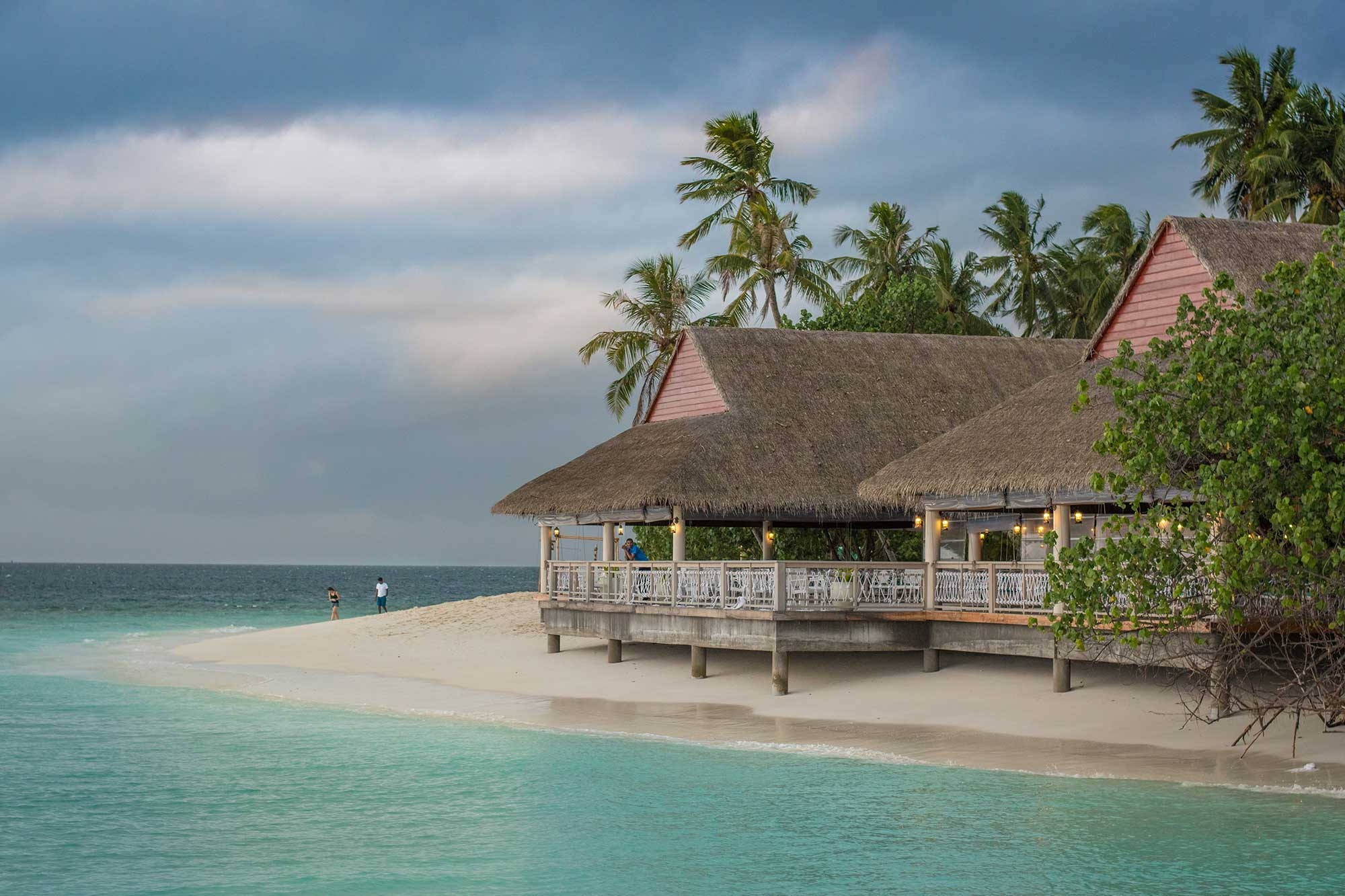 Beach House 桌布 壁紙 — 寧靜的熱帶海灘場景，茅草屋頂的水上平房、棕櫚樹和碧綠的海水，天空多雲。