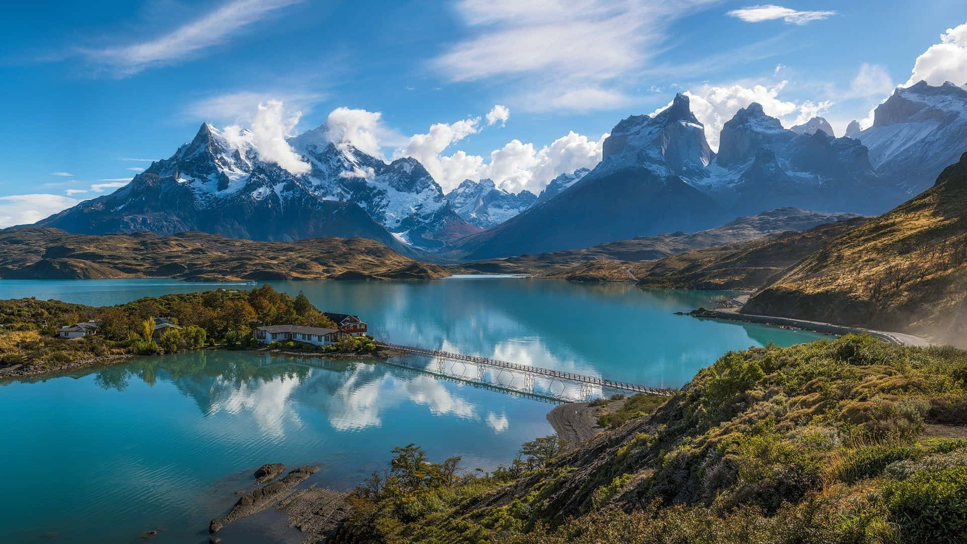 Patagonia 桌布 壁紙 — 寧靜的碧綠湖面映照著白雪覆蓋的山峰和蓬鬆的雲朵，在明亮的藍天下，一座木橋連接兩岸。