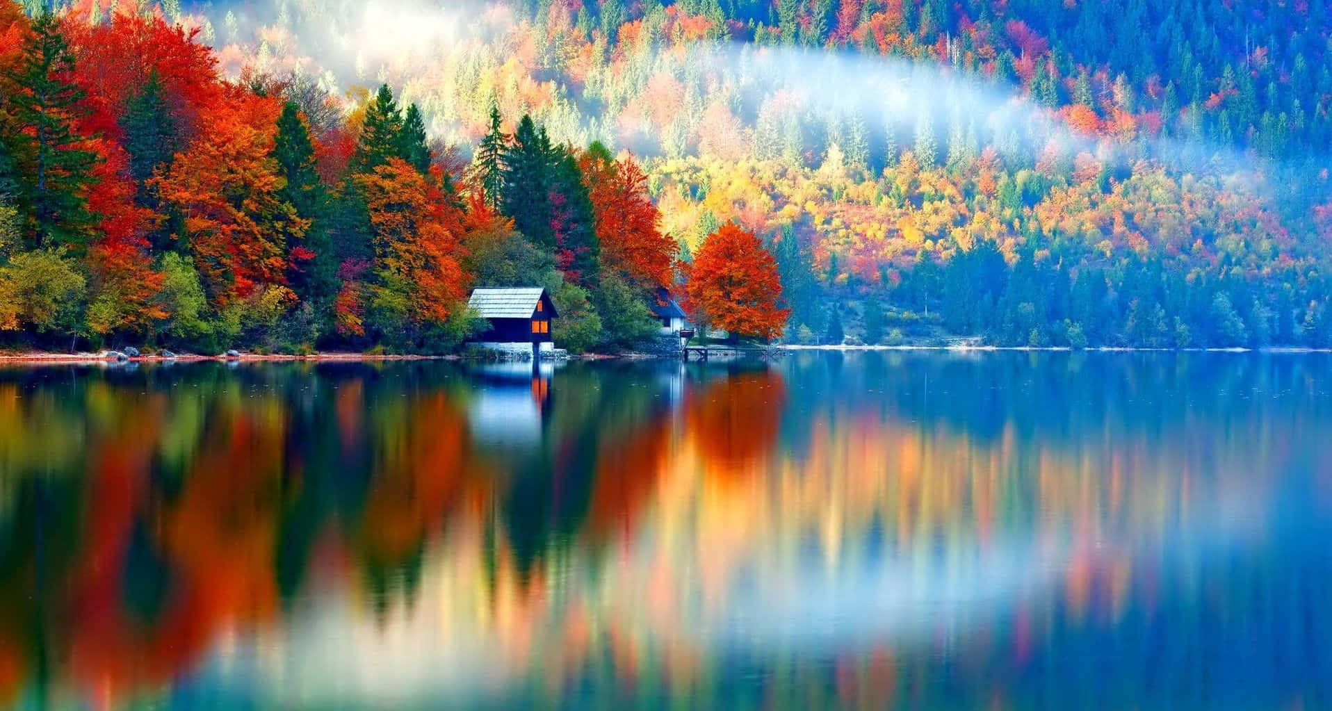 Fall Colors 桌布 壁紙 — 寧靜的秋日湖面倒映著火紅、橙黃和金黃的樹木，其間有一座小木屋，坐落在霧氣繚繞的山丘下。
