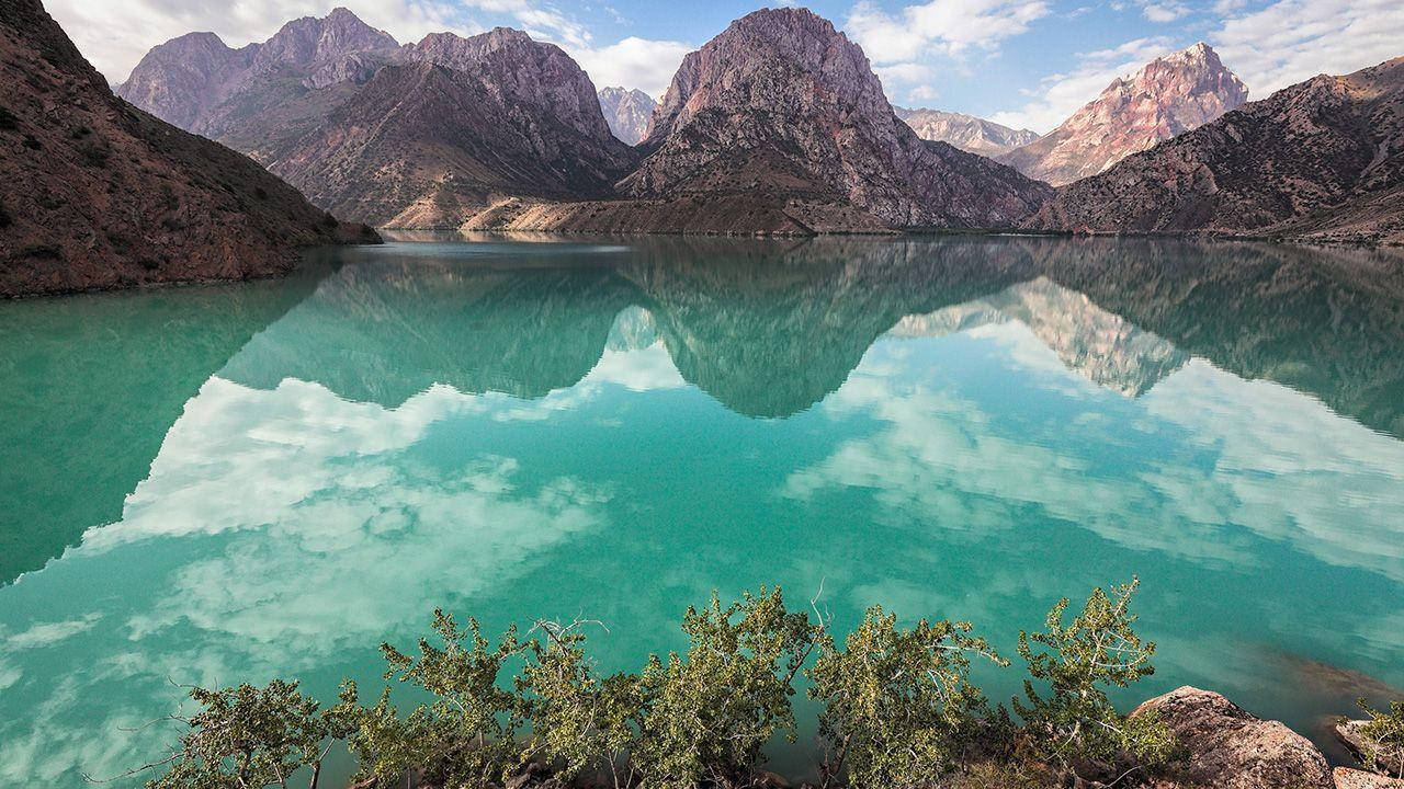 Tajikistan 桌布 壁紙 — 寧靜的綠松石湖面倒映著崎嶇的山脈和陰雲密布的天空，前景點綴著綠色灌木。