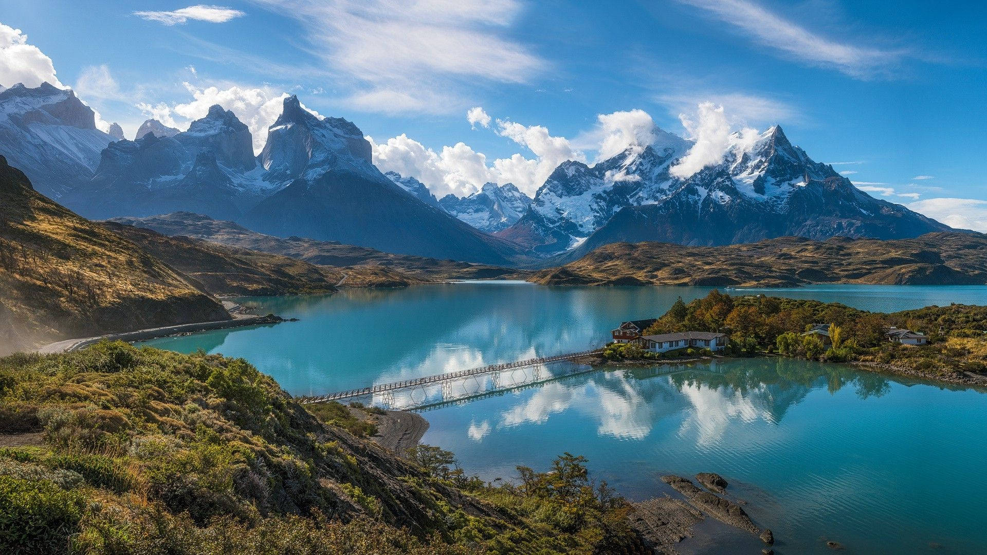 Patagonia 桌布 壁紙 — 寧靜的綠松石湖面倒映著積雪的山峰和蓬鬆的雲朵，在明亮的藍天下，一座木橋連接兩岸。
