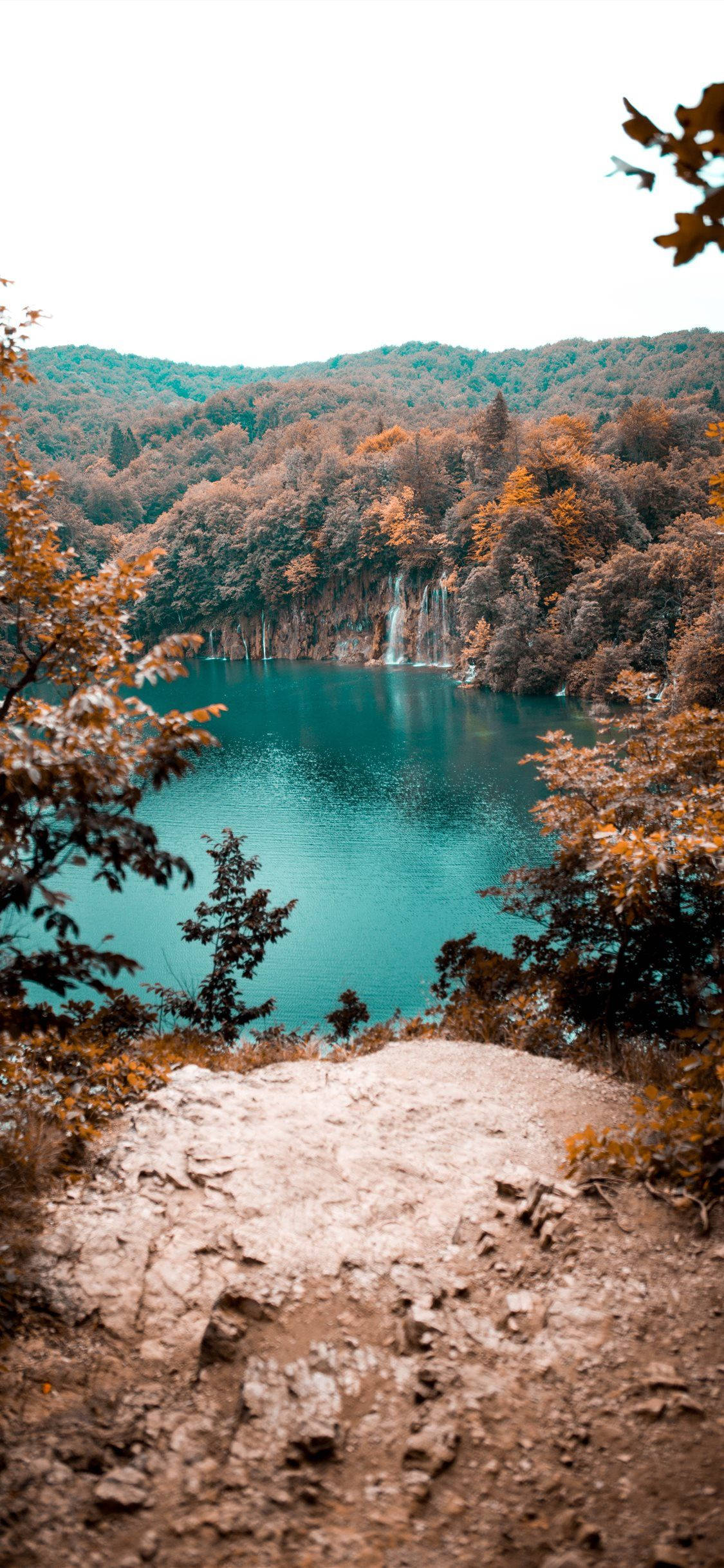 View 桌布 壁紙 — 寧靜的綠松石色湖泊隱藏在秋日樹林中，瀑布傾瀉入平靜的水面，四周環繞著金色的樹葉。