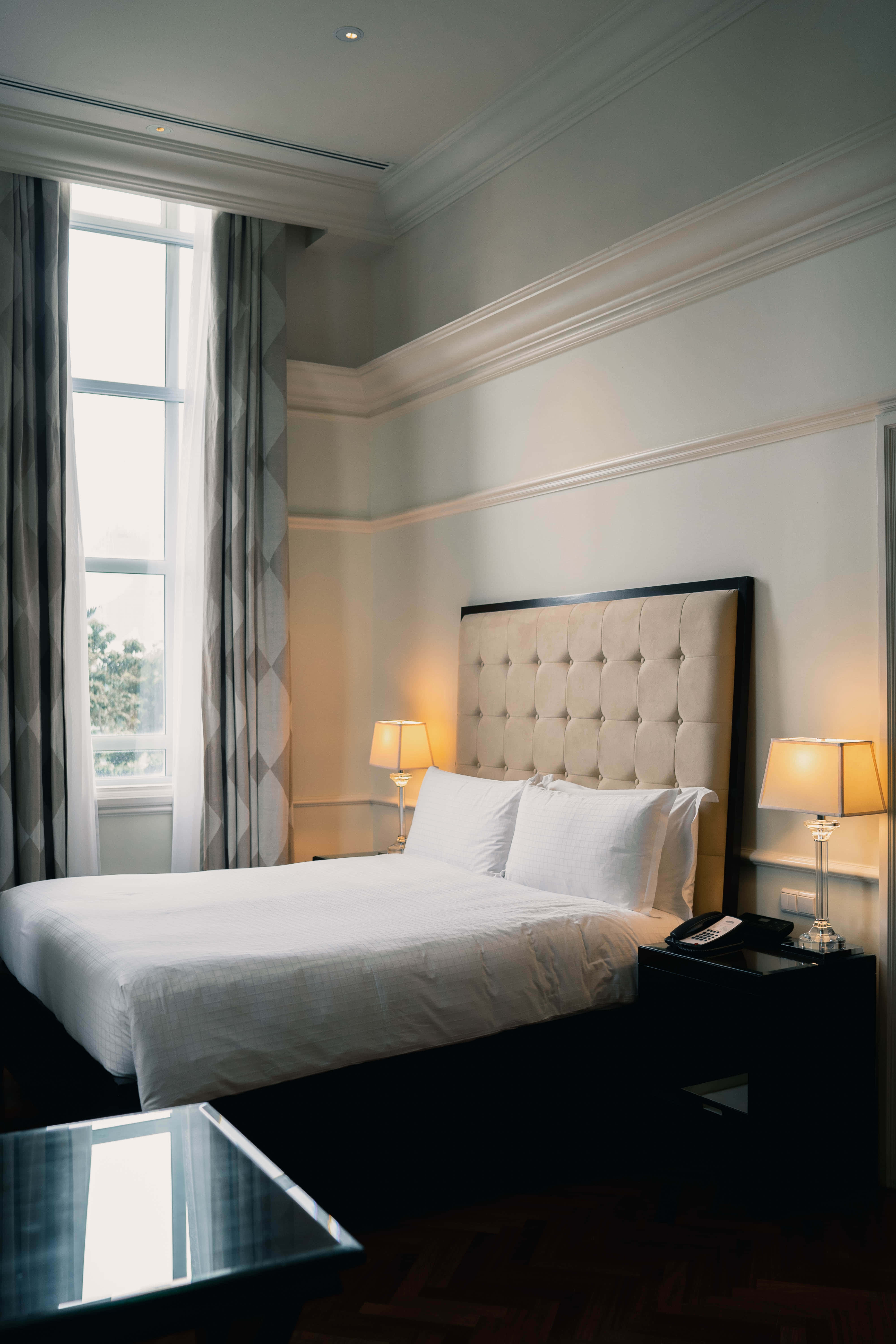 Hotel Room 桌布 壁紙 — 寧靜的飯店臥室，配有簇絨奶油色床頭板、白色床單和散發柔和光線的溫暖床頭燈。