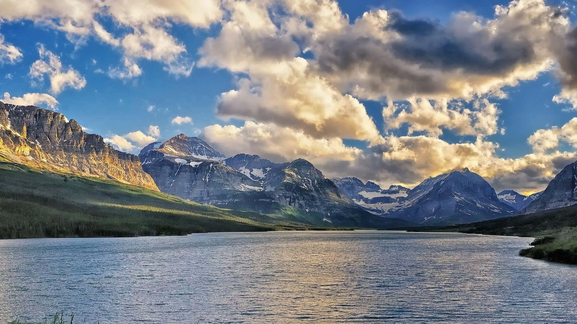 Montana 桌布 壁紙 — 寧靜的高山湖泊在日落時分戲劇性的天空下，映照著白雪覆蓋的山峰，天空中有金色的雲彩。