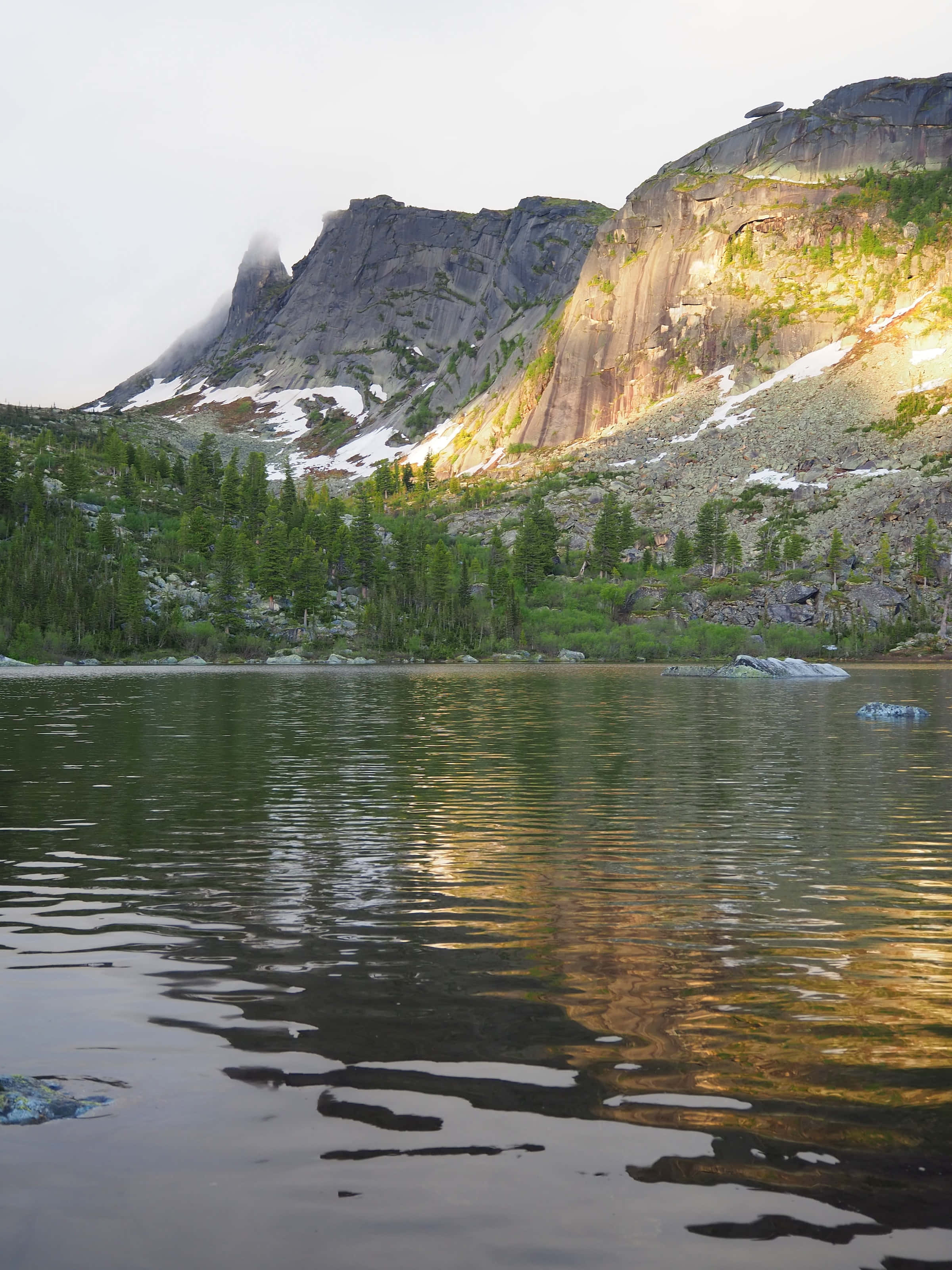 Lake 桌布 壁紙 — 寧靜的高山湖泊映照著崎嶇的山峰，沐浴在金色的夕陽餘暉中，殘留的雪斑和常綠樹點綴其間。