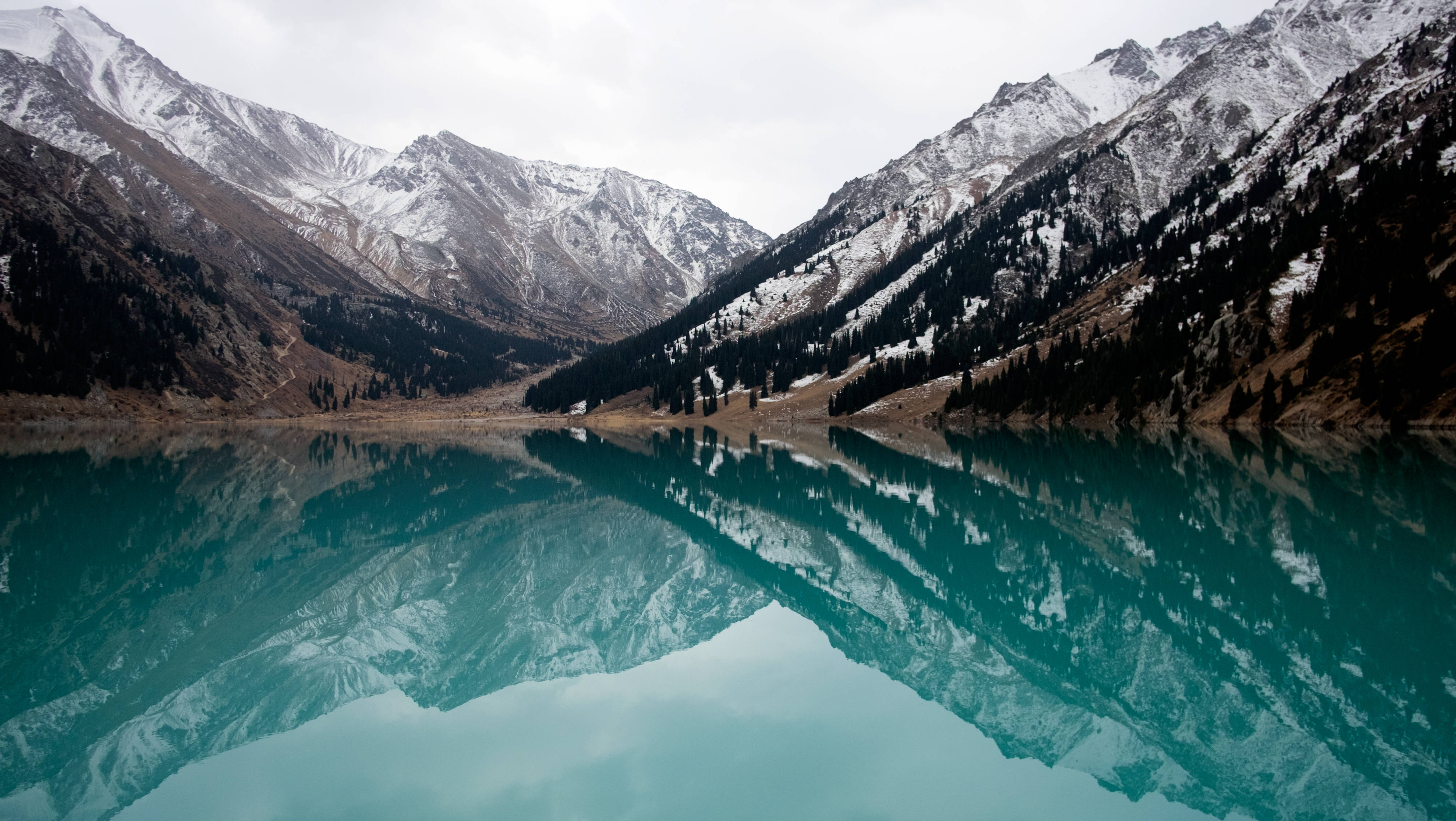 Kazakhstan 桌布 壁紙 — 寧靜的高山湖泊映照著白雪覆蓋的山峰，天空多雲，湖水呈深藍綠色，周圍是常綠森林。
