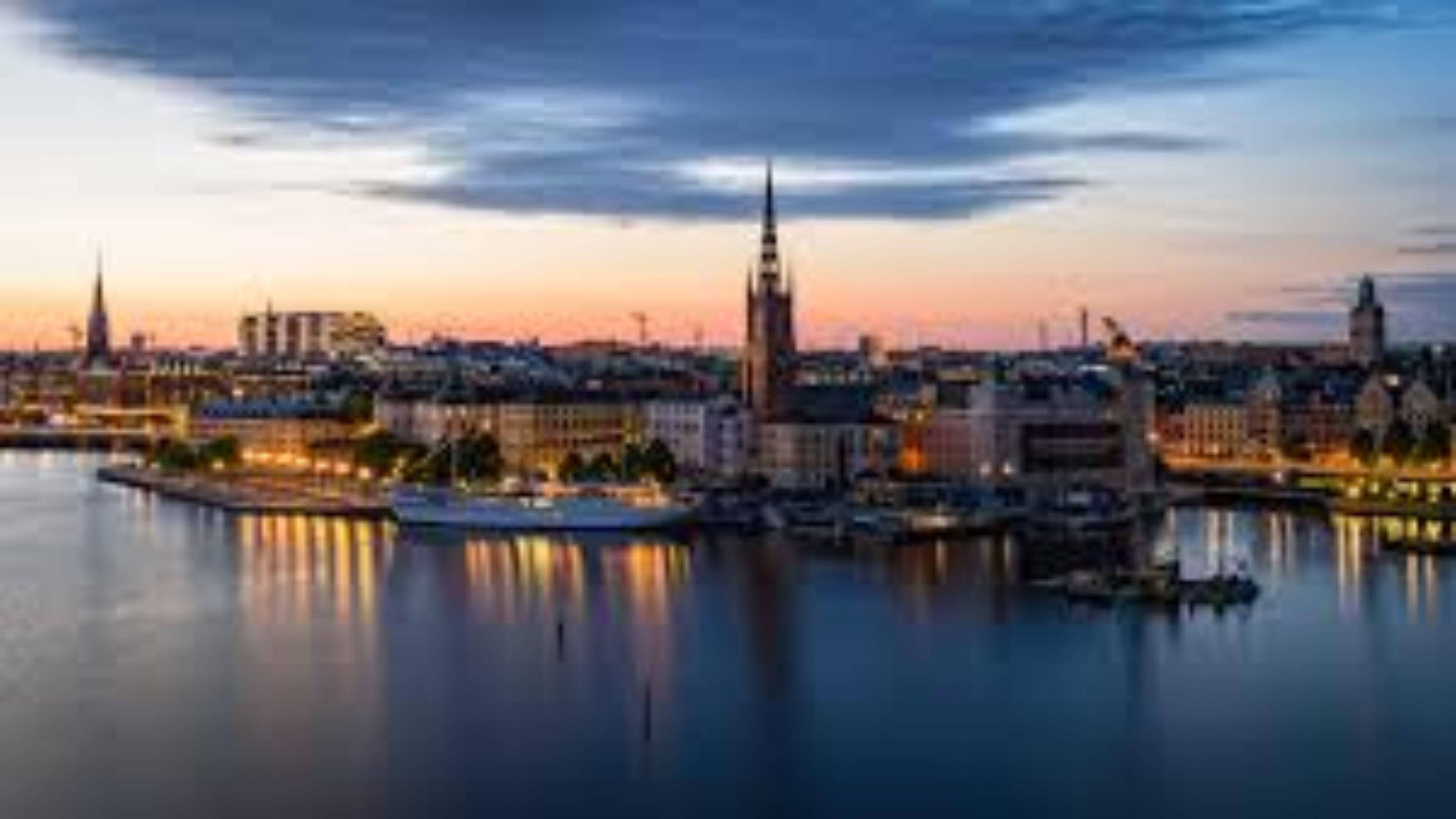 Sweden 桌布 壁紙 — 寧靜的黃昏城市景觀，歷史悠久的尖塔和發光的燈光倒映在平靜的水面上，天空呈現柔和的橙色和藍色。