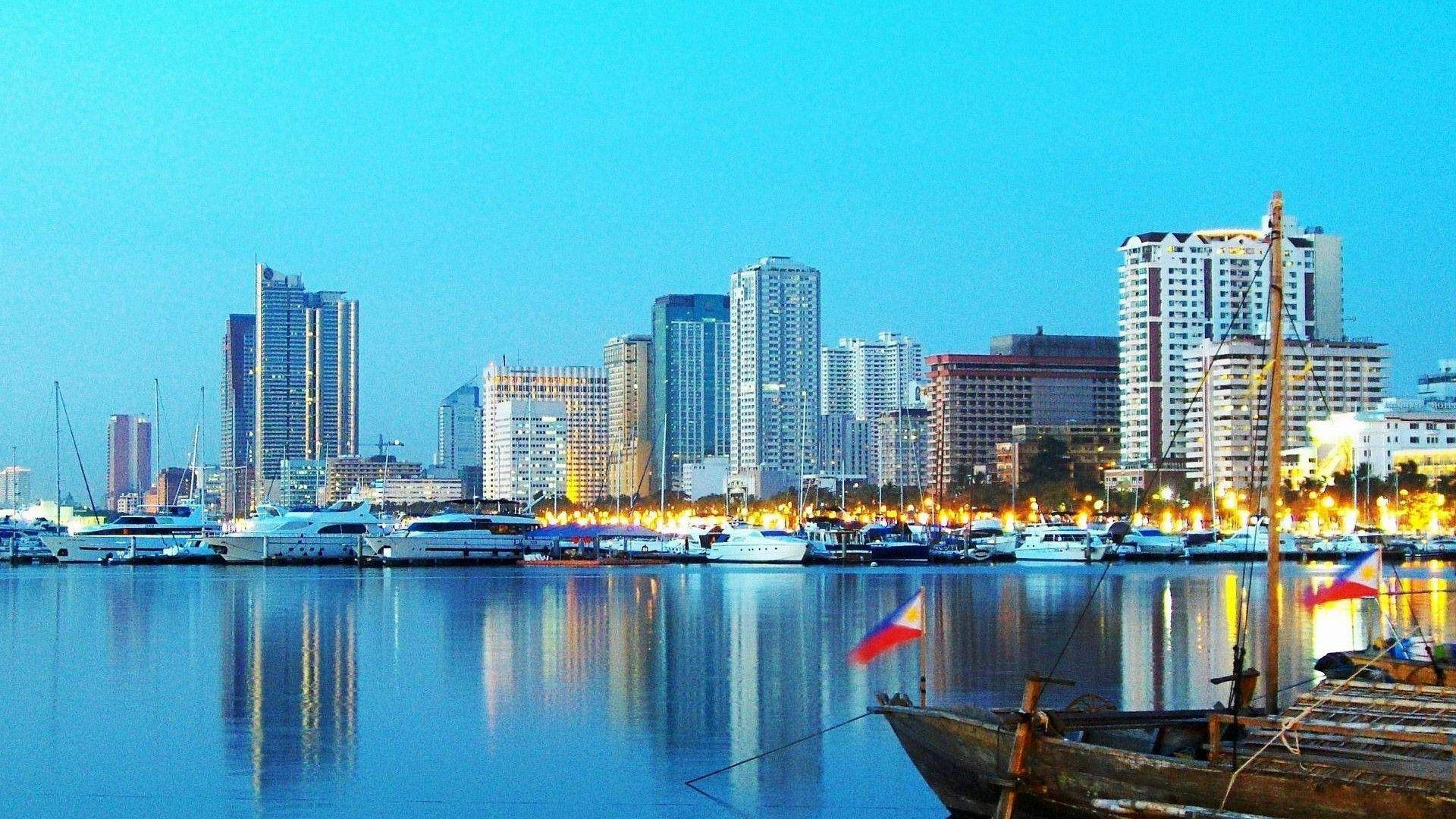 馬尼拉 桌布 壁紙 — 寧靜的黃昏城市景觀，現代摩天大樓倒映在平靜的水面上，旁邊是傳統船隻。