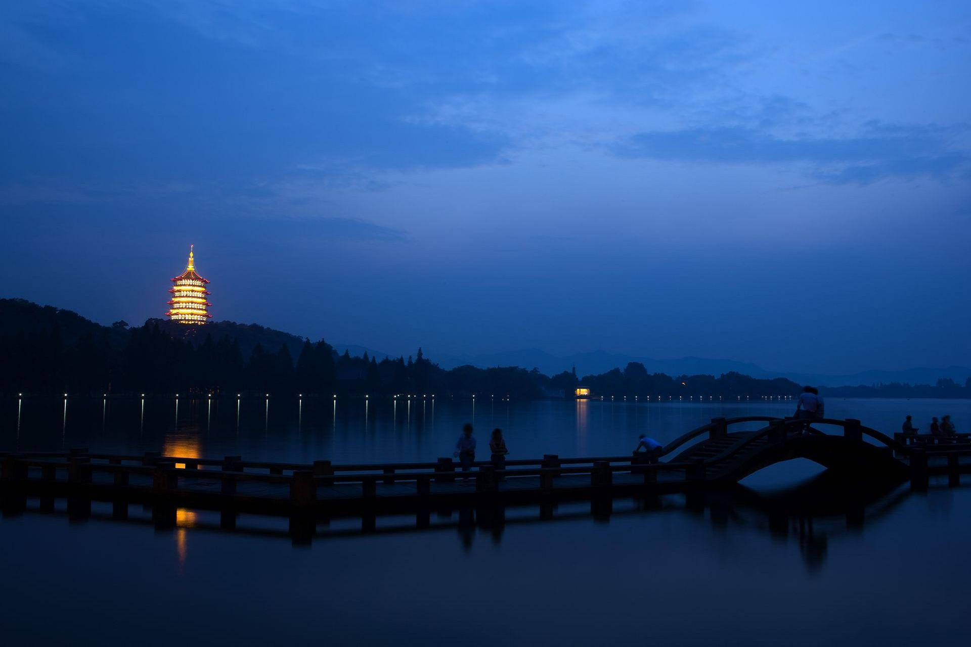 Hangzhou 桌布 壁紙 — 寧靜的黃昏場景，山丘上發光的寶塔倒映在平靜的水面上，木橋上的人影剪影。