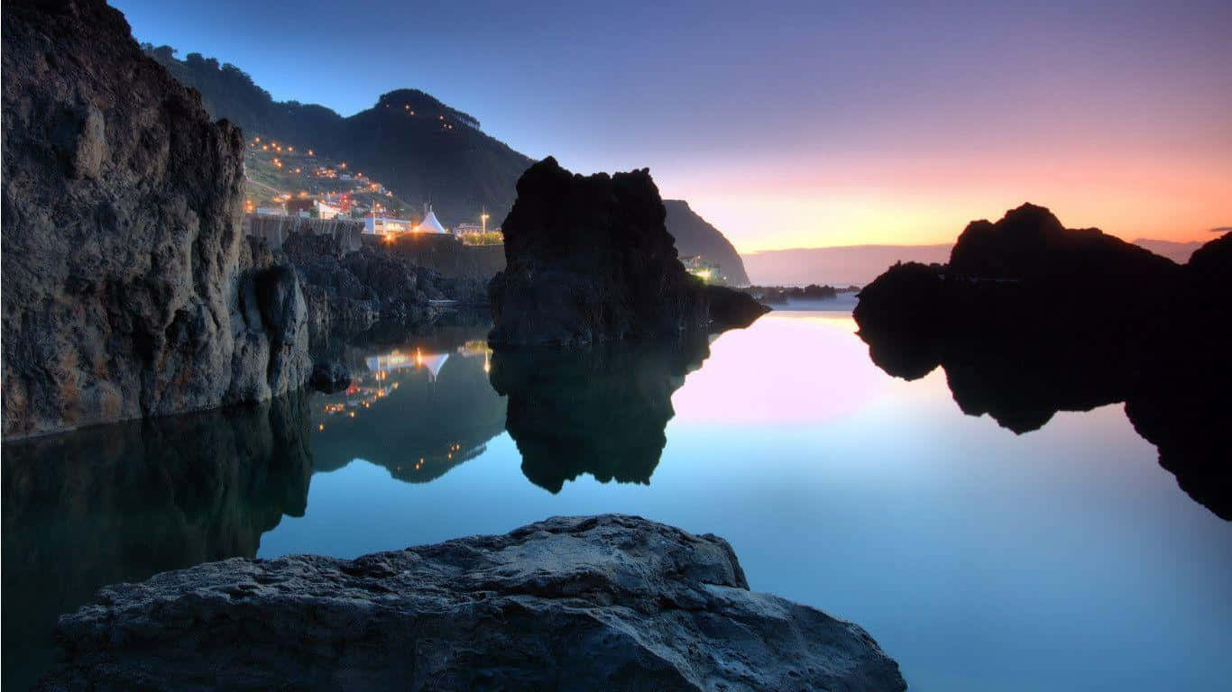 Nature Landscape 桌布 壁紙 — 寧靜的黃昏場景，村莊燈光閃爍，倒映在平靜的水面上，周圍環繞著深色的火山岩層。