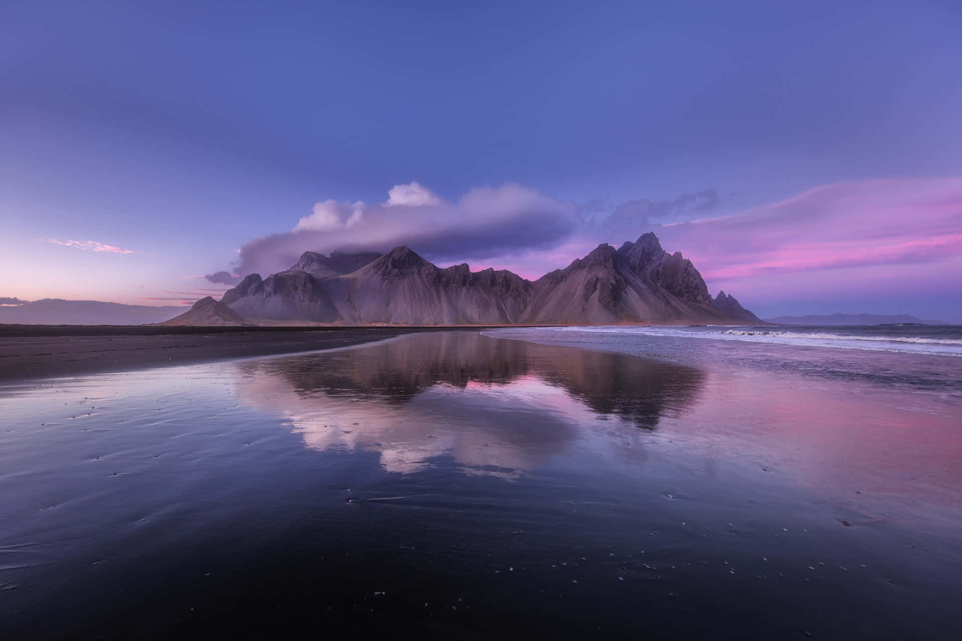 Iceland Desktop 桌布 壁紙 — 寧靜的黃昏場景，鋸齒狀的山脈在紫色和粉紅色的天空下倒映在濕潤的黑沙上。
