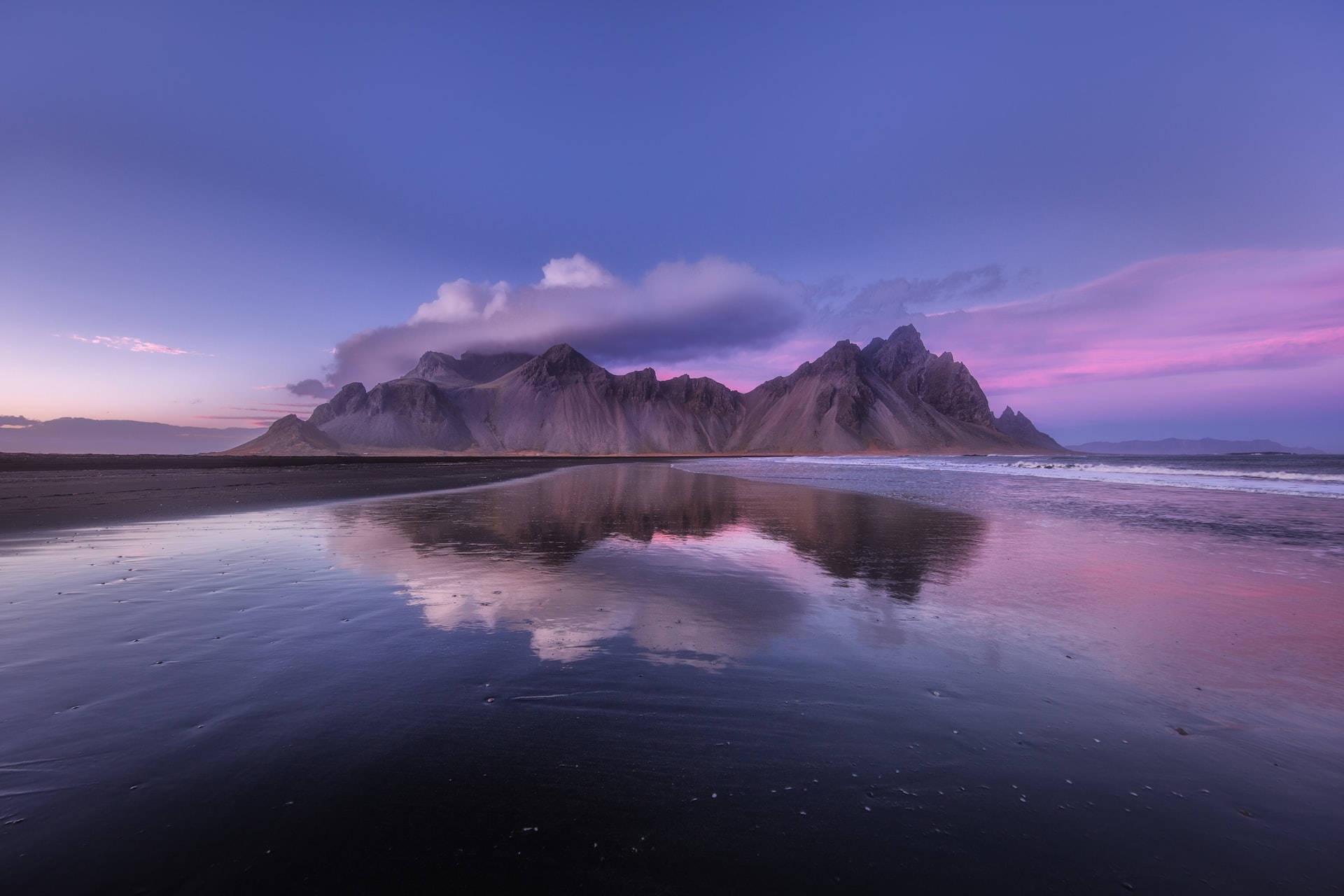 Mountain Landscape 桌布 壁紙 — 寧靜的黃昏場景，鋸齒狀的山脈在紫色和粉紅色的天空下倒映在平靜的黑沙灘上。