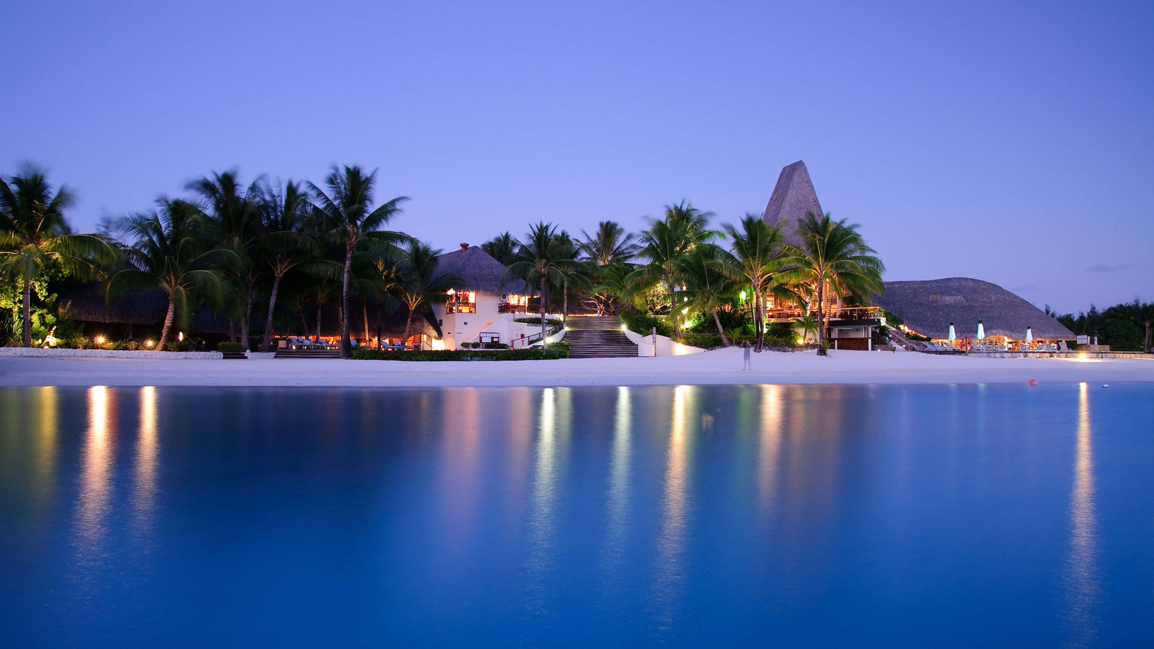 Bora Bora 桌布 壁紙 — 寧靜的黃昏度假村，棕櫚樹和茅草屋頂倒映在平靜的藍色海水中。