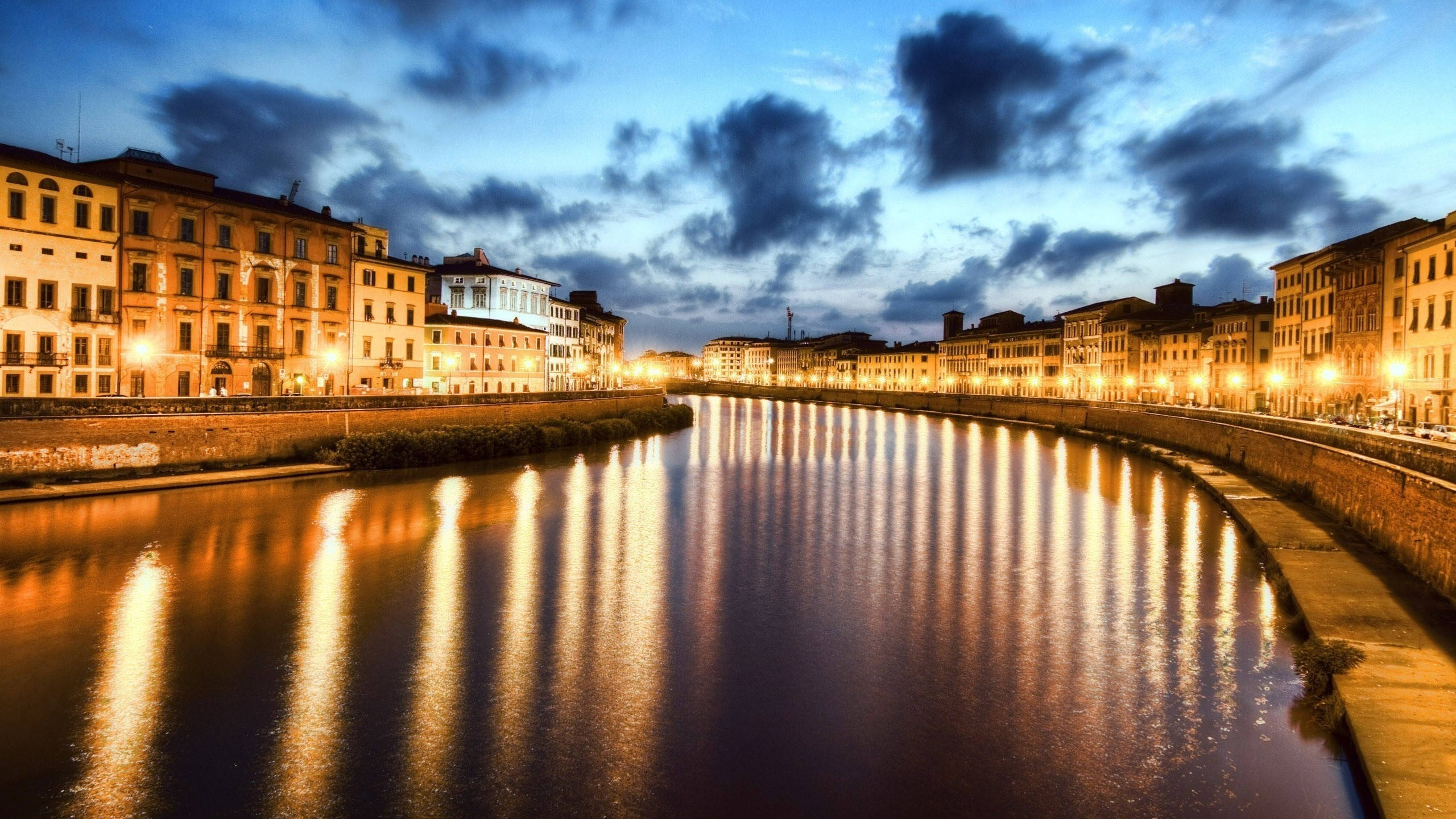 Pisa 桌布 壁紙 — 寧靜的黃昏河流映照著金色的街燈和歷史建築，背景是戲劇性的藍天。