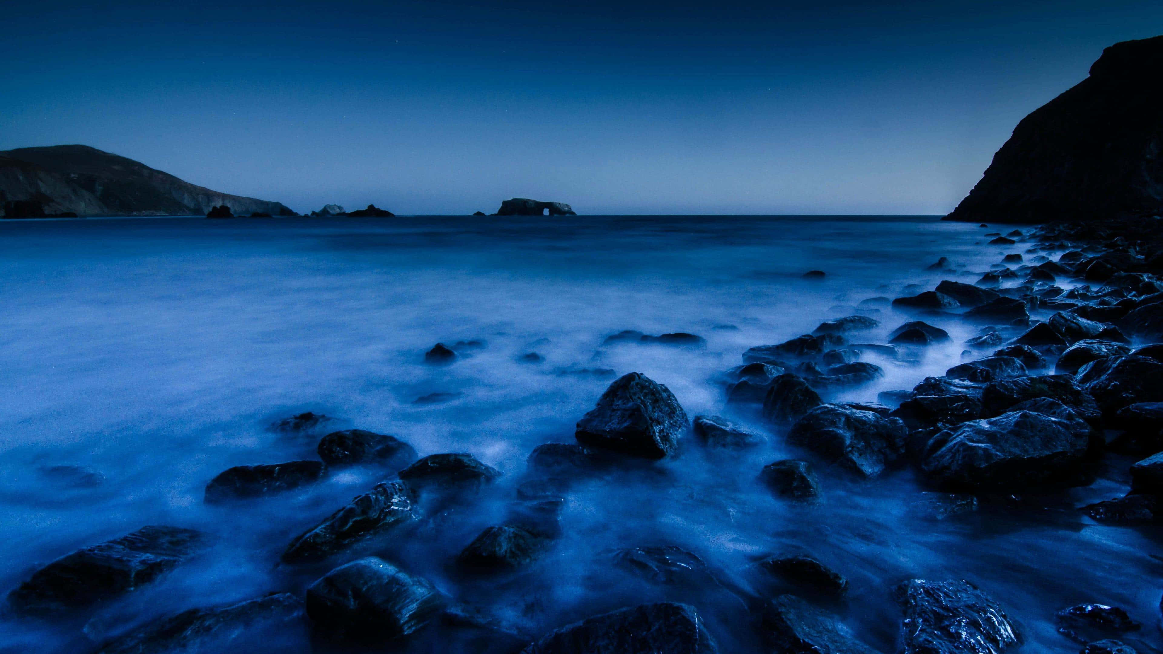 水 桌布 壁紙 — 寧靜的黃昏海景，平滑的藍色水流過深色岩石，天空深藍。
