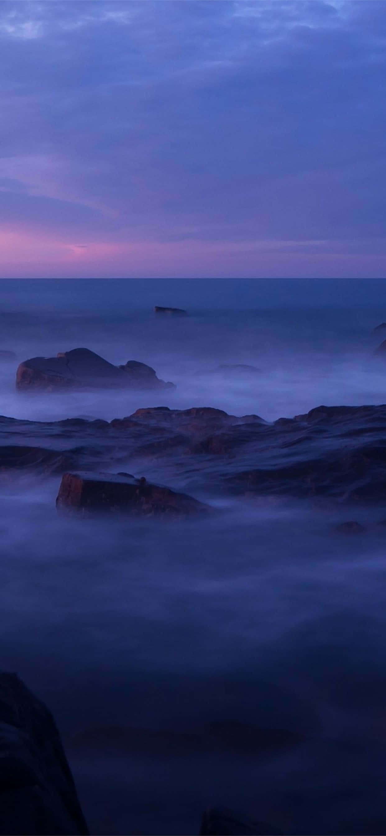 Calm Aesthetic 桌布 壁紙 — 寧靜的黃昏海景，霧氣朦朧的海浪拍打著深色岩石，天空呈現紫色和粉紅色。