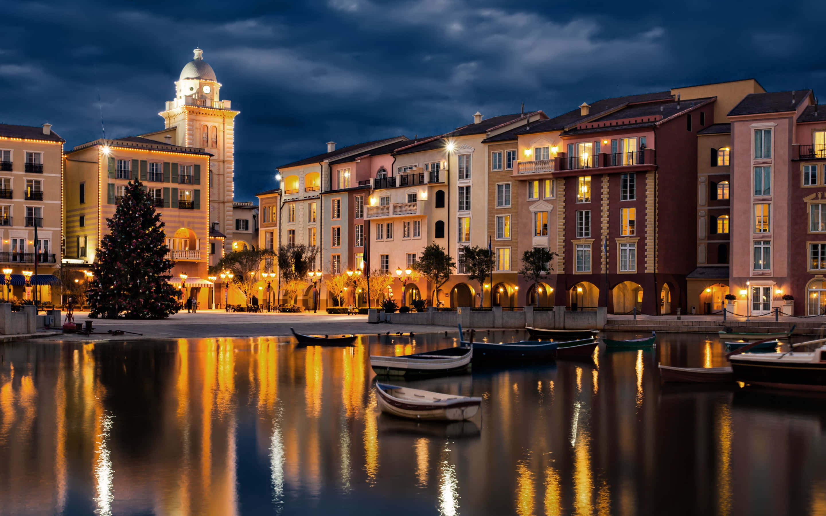 Orlando 桌布 壁紙 — 寧靜的黃昏海濱，溫暖燈光照亮的歐式建築倒映在平靜的水面上，旁邊停泊著船隻和裝飾好的聖誕樹。