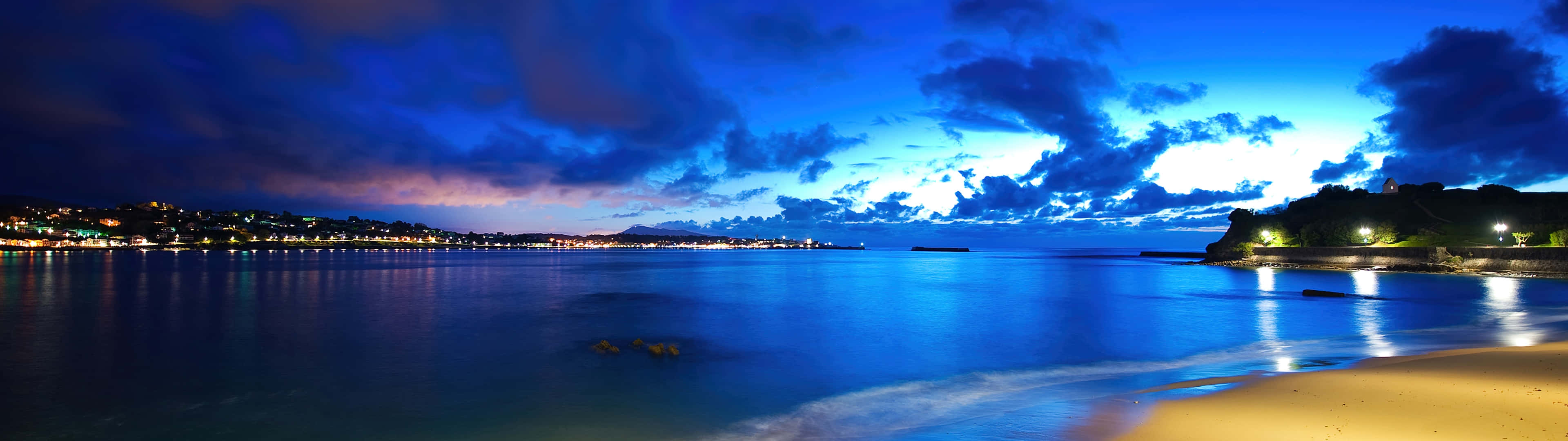 夜晚 桌布 壁紙 — 寧靜的黃昏海灘全景，城市燈光在平靜的水面上閃爍，映襯著戲劇性的藍天。