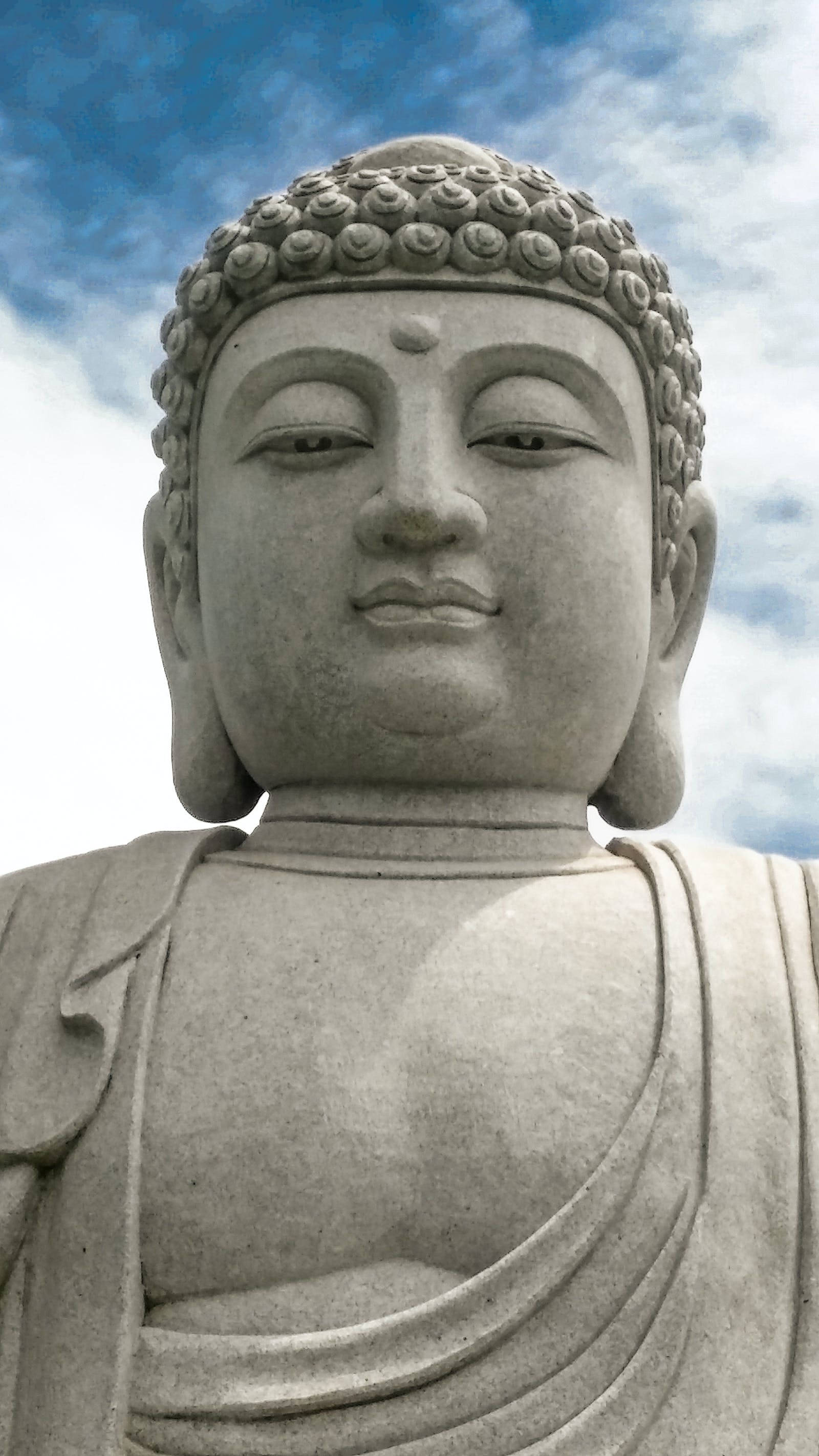 Gautam Buddha 桌布 壁紙 — 寧靜石佛像特寫，捲髮，表情平靜，背景是明亮藍天和縷縷白雲。