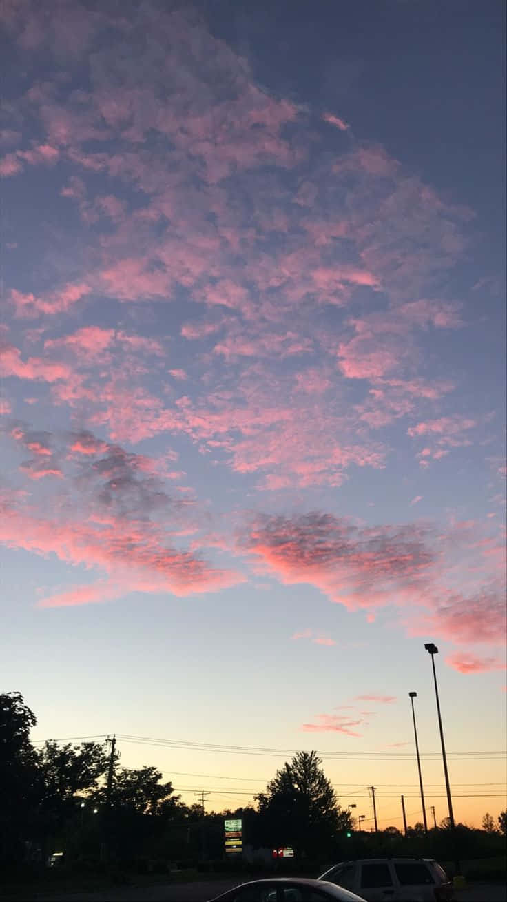 雲朵 桌布 壁紙 — 寧靜路邊停車場上空，鮮豔的粉紅和紫色日落雲景，襯托出樹木和汽車的剪影。