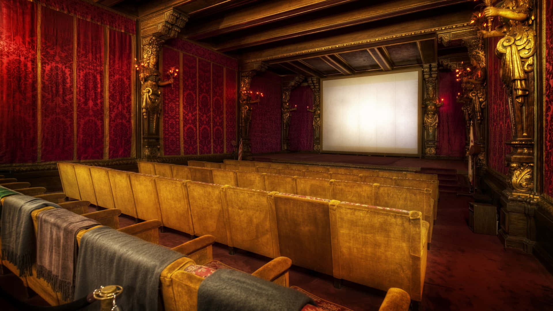 Home Cinema 桌布 壁紙 — 寬敞的復古電影院，配有豪華紅色天鵝絨牆壁、金色華麗細節，以及一排排磨損的天鵝絨座椅，面向明亮的空白螢幕。