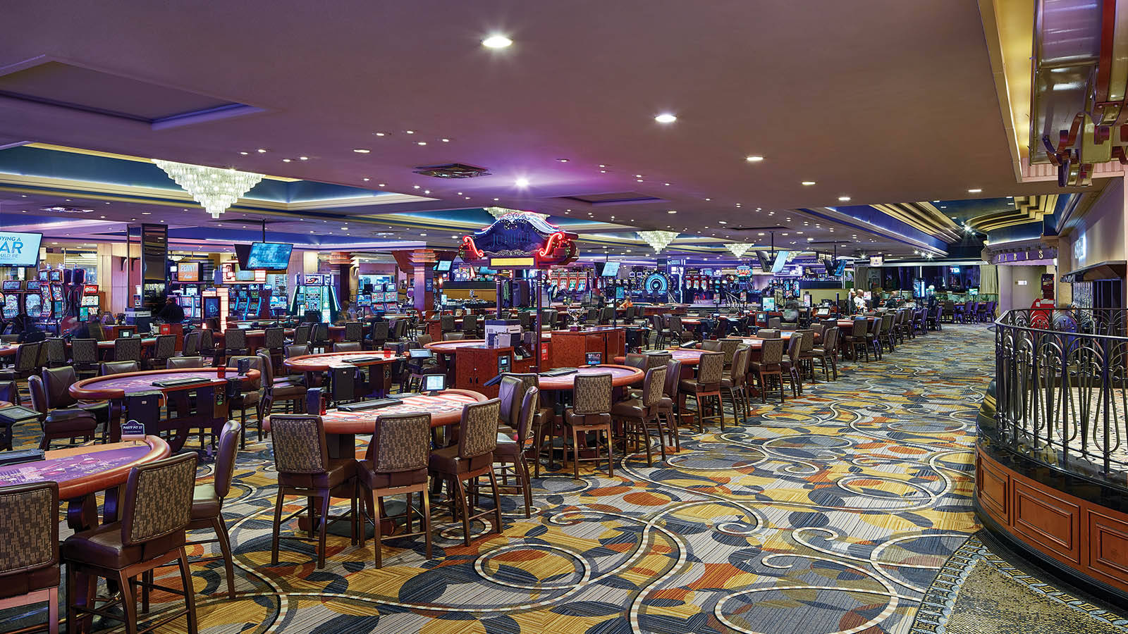 Casino 桌布 壁紙 — 寬敞的賭場地面，擺滿了撲克桌、老虎機和色彩鮮豔的圖案地毯，在溫暖的環境燈光下。