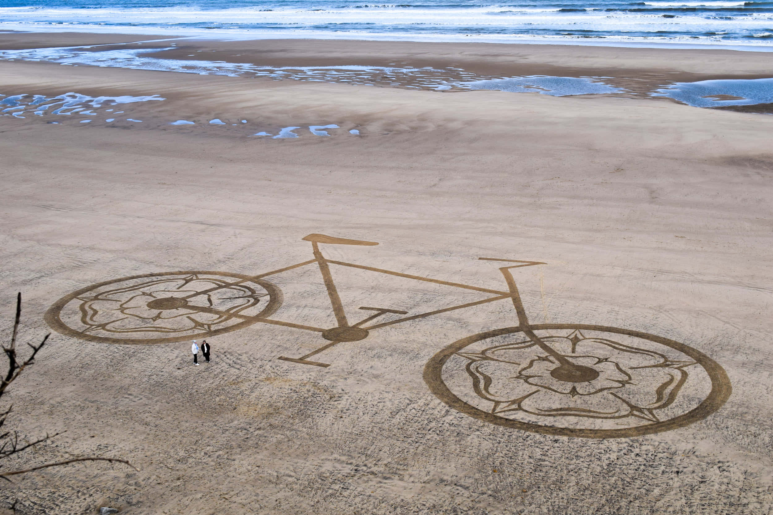 Sand Art 桌布 壁紙 — 寬闊沙灘上巨型腳踏車沙雕的鳥瞰圖，兩人站在其旁邊，靠近海岸線。
