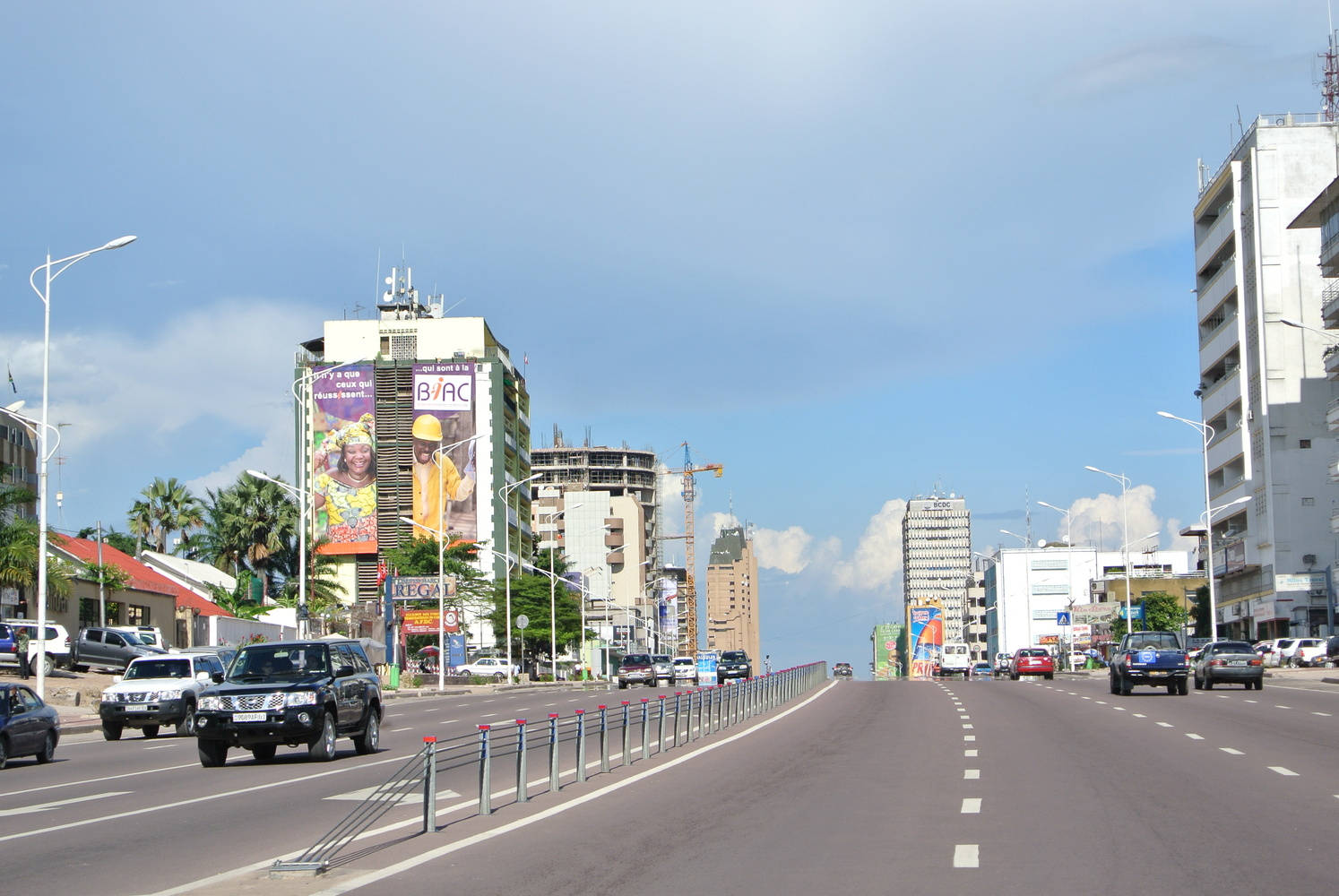 Kinshasa 桌布 壁紙 — 寬闊的城市街道，有汽車和高樓大廈，在明亮的藍天下有零星雲朵。