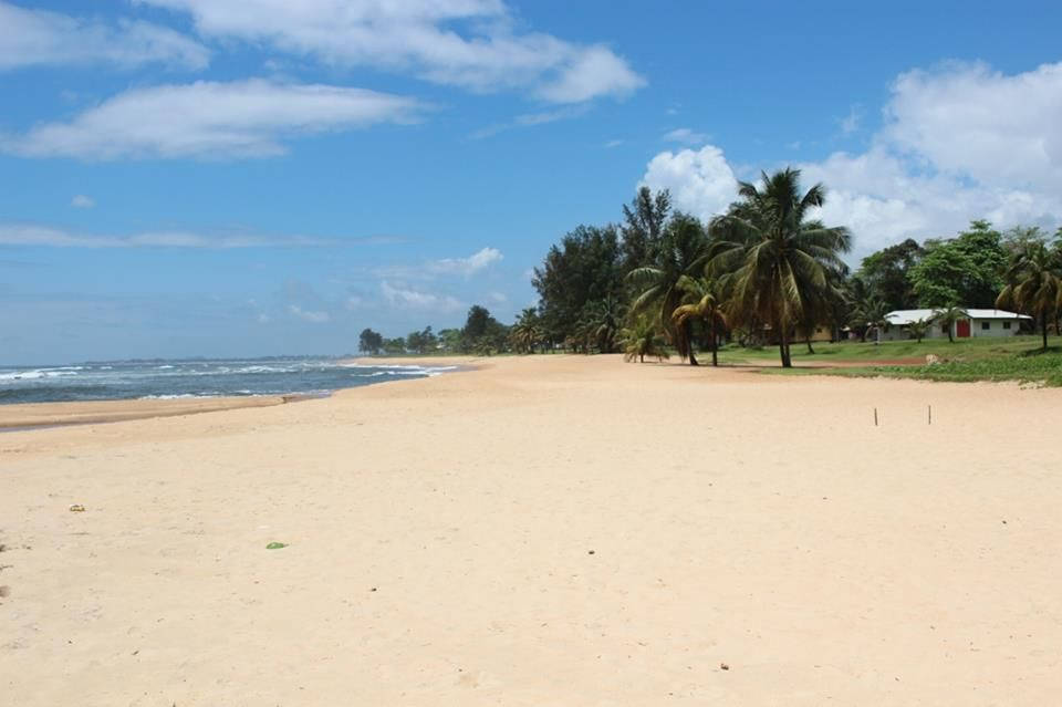 Liberia 桌布 壁紙 — 寬闊的沙灘在明亮的藍天下延伸，有蓬鬆的雲朵，海浪在棕櫚樹和遠處建築物旁翻滾。