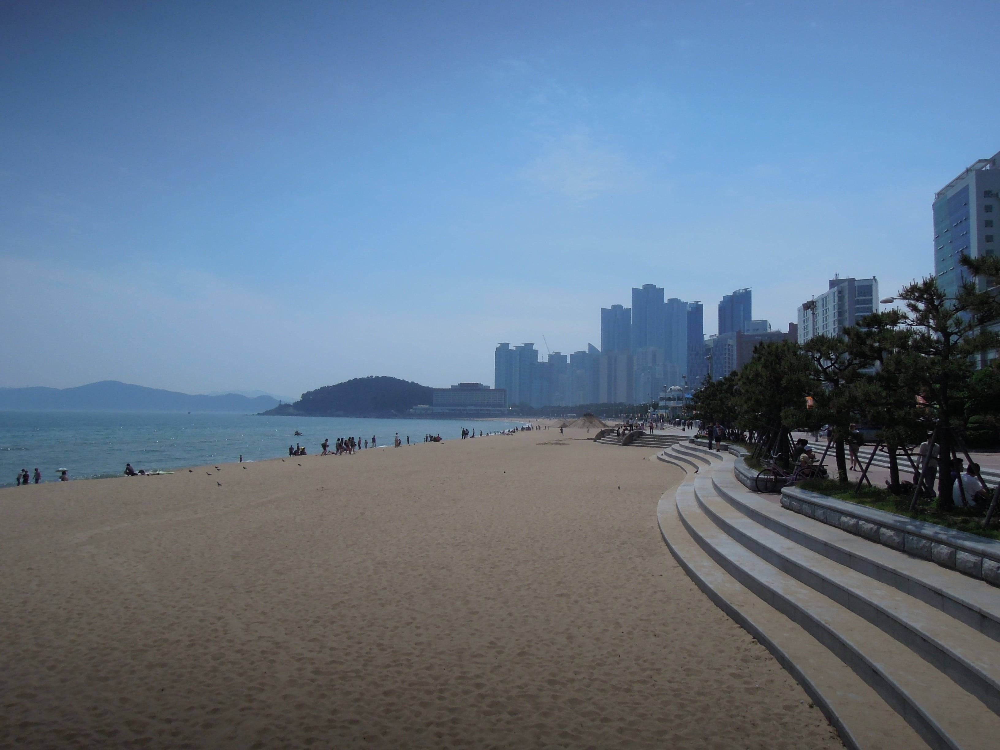 Busan 桌布 壁紙 — 寬闊的沙灘在晴朗的藍天下延伸至現代城市天際線，人們享受著海岸。
