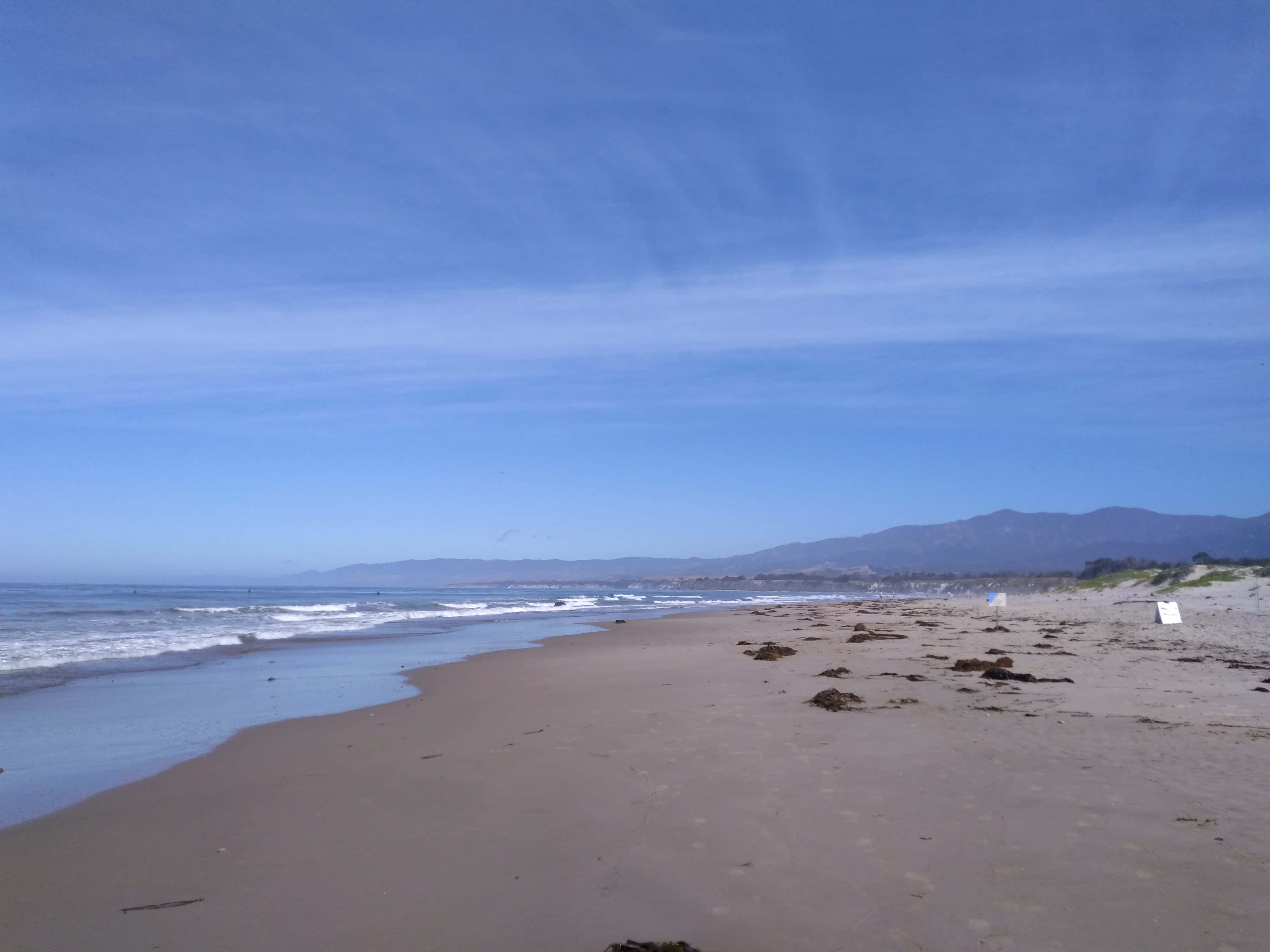 Beach Sand 桌布 壁紙 — 寬闊的沙灘在清澈的藍天下延伸，溫柔的海浪拍打著岸邊，遠山在地平線上若隱若現。