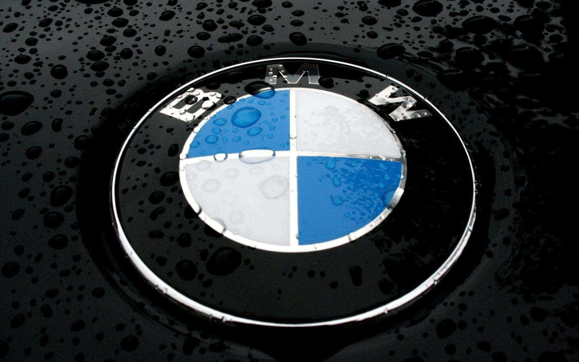 Bmw Logo 桌布 壁紙 — 寶馬標誌特寫，位於濕透的黑色引擎蓋上，雨滴在明亮燈光下閃閃發光。