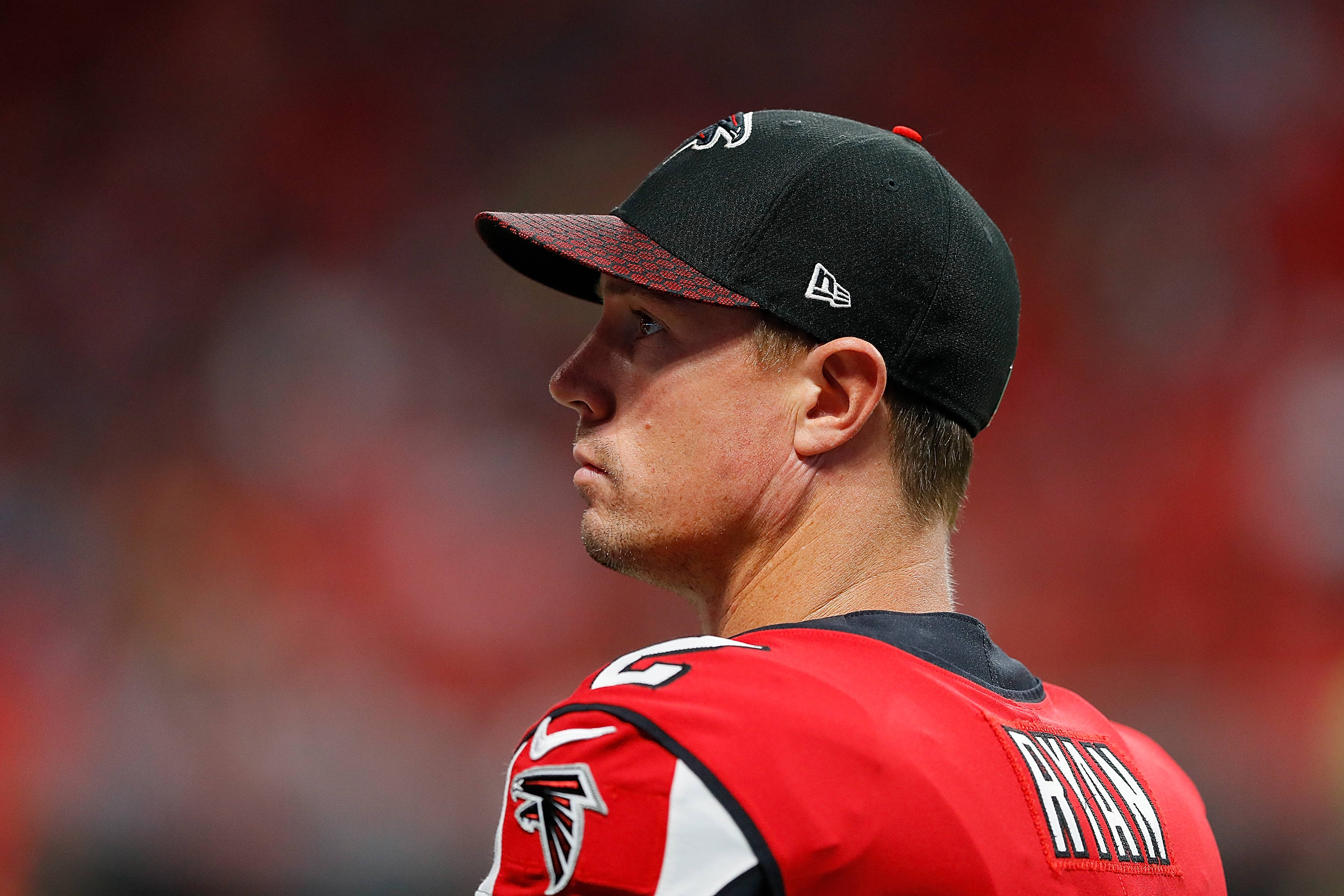 Matt Ryan 桌布 壁紙 — 專注的亞特蘭大獵鷹美式足球員身穿紅色球衣和黑色帽子，背景是模糊的體育場人群的特寫側面照。