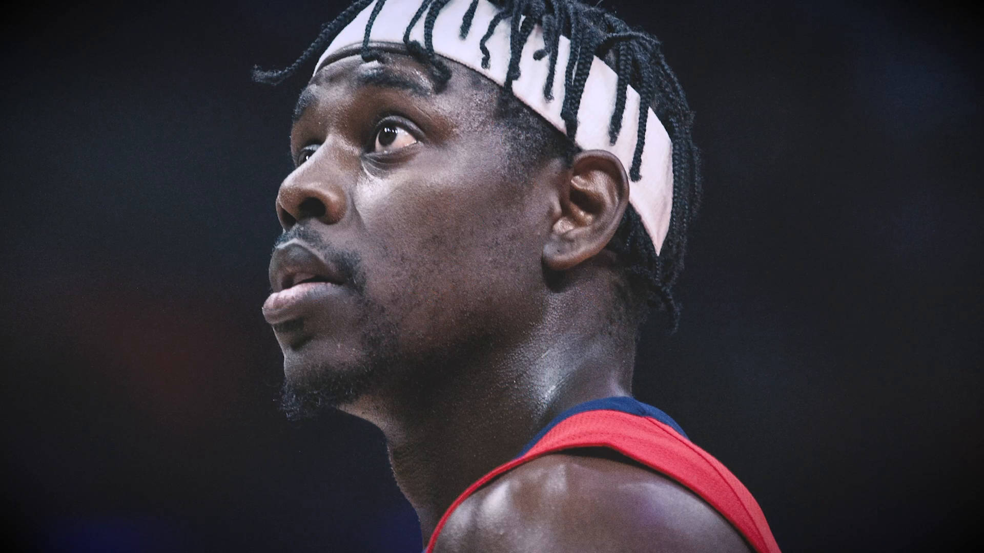 Jrue Holiday 桌布 壁紙 — 專注的籃球運動員特寫，戴著白色頭帶，黑色辮子頭，背景深色。
