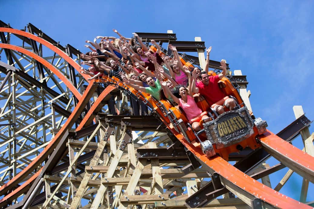 Six Flags 桌布 壁紙 — 尋求刺激的人在晴朗的藍天下，在充滿活力的橙色木製雲霄飛車上倒掛飛馳，歡呼尖叫。