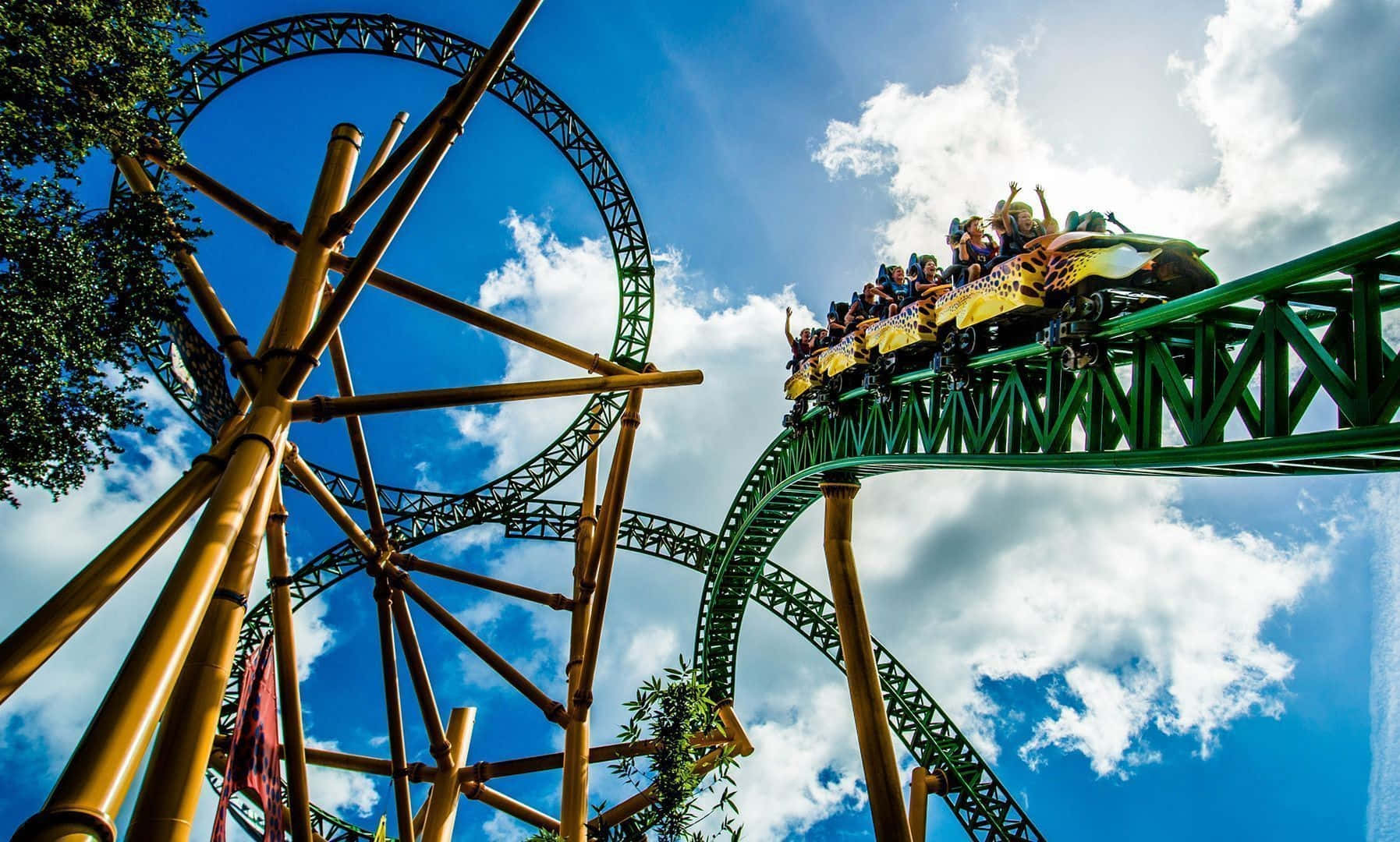 Roller Coaster 桌布 壁紙 — 尋求刺激的人在歡呼聲中，一輛黃色雲霄飛車在明亮的藍天和 fluffy 白雲背景下，沿著蜿蜒的綠色軌道疾馳。