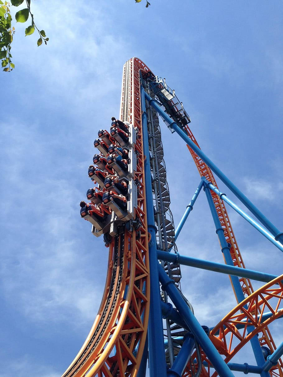 Roller Coaster 桌布 壁紙 — 尋求刺激的人在雲霄飛車沿著高聳的橙色和藍色軌道上升，映襯著明亮的藍天時尖叫。