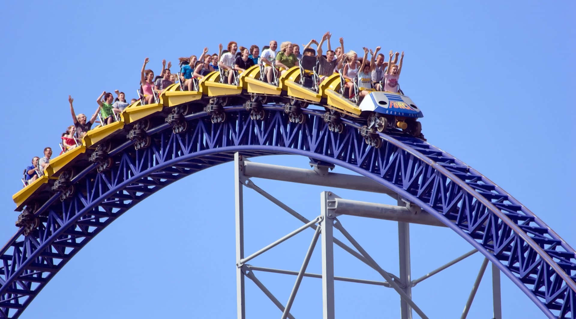 Roller Coaster 桌布 壁紙 — 尋求刺激的人在黃色雲霄飛車車廂高聳入蔚藍天空時歡呼尖叫。