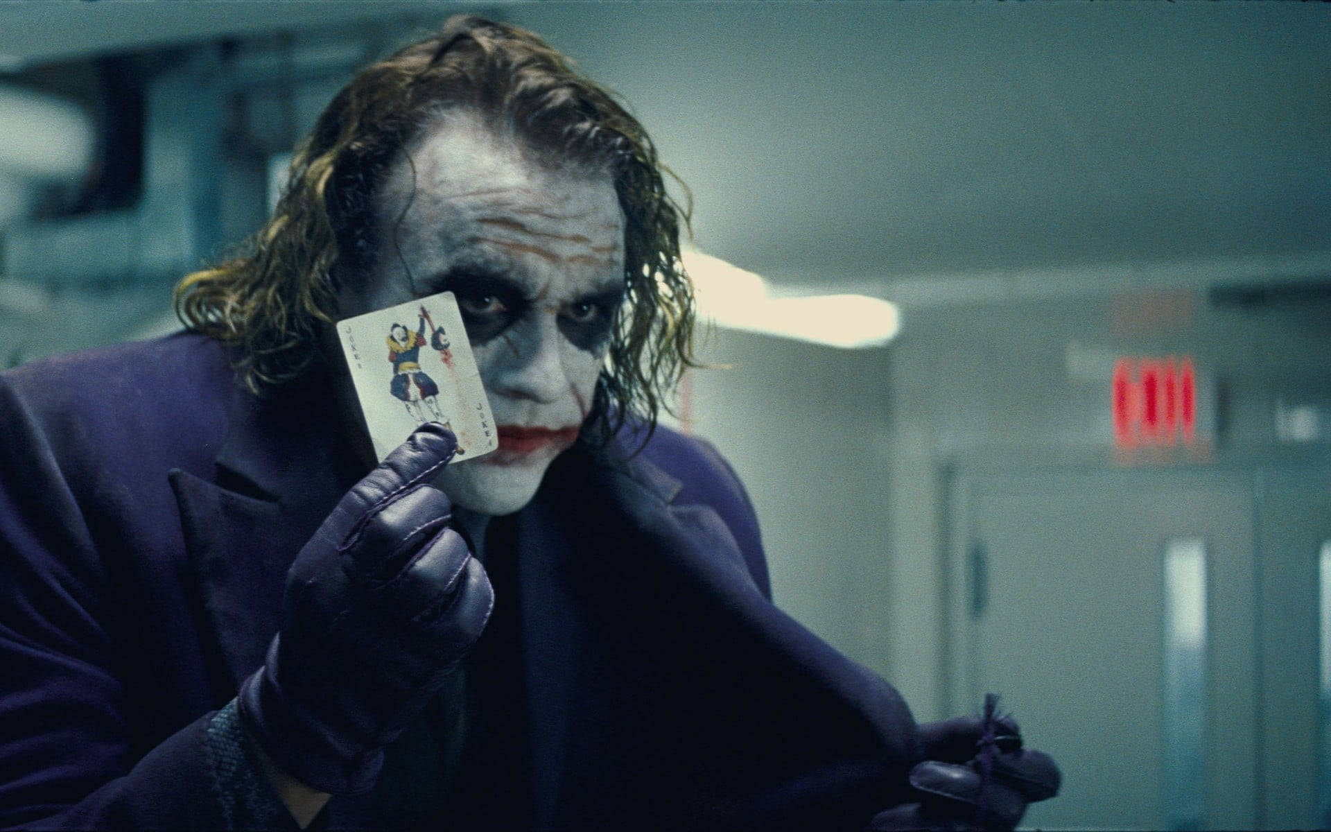 Heath Ledger Joker 桌布 壁紙 — 小丑在昏暗的走廊裡舉起一張撲克牌，露出邪惡的笑容和模糊的化妝。