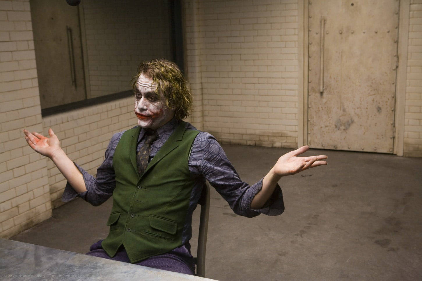 Heath Ledger Joker 桌布 壁紙 — 小丑坐在一個簡陋的磚房裡，雙臂張開，穿著他標誌性的綠色背心和白色臉部彩繪。