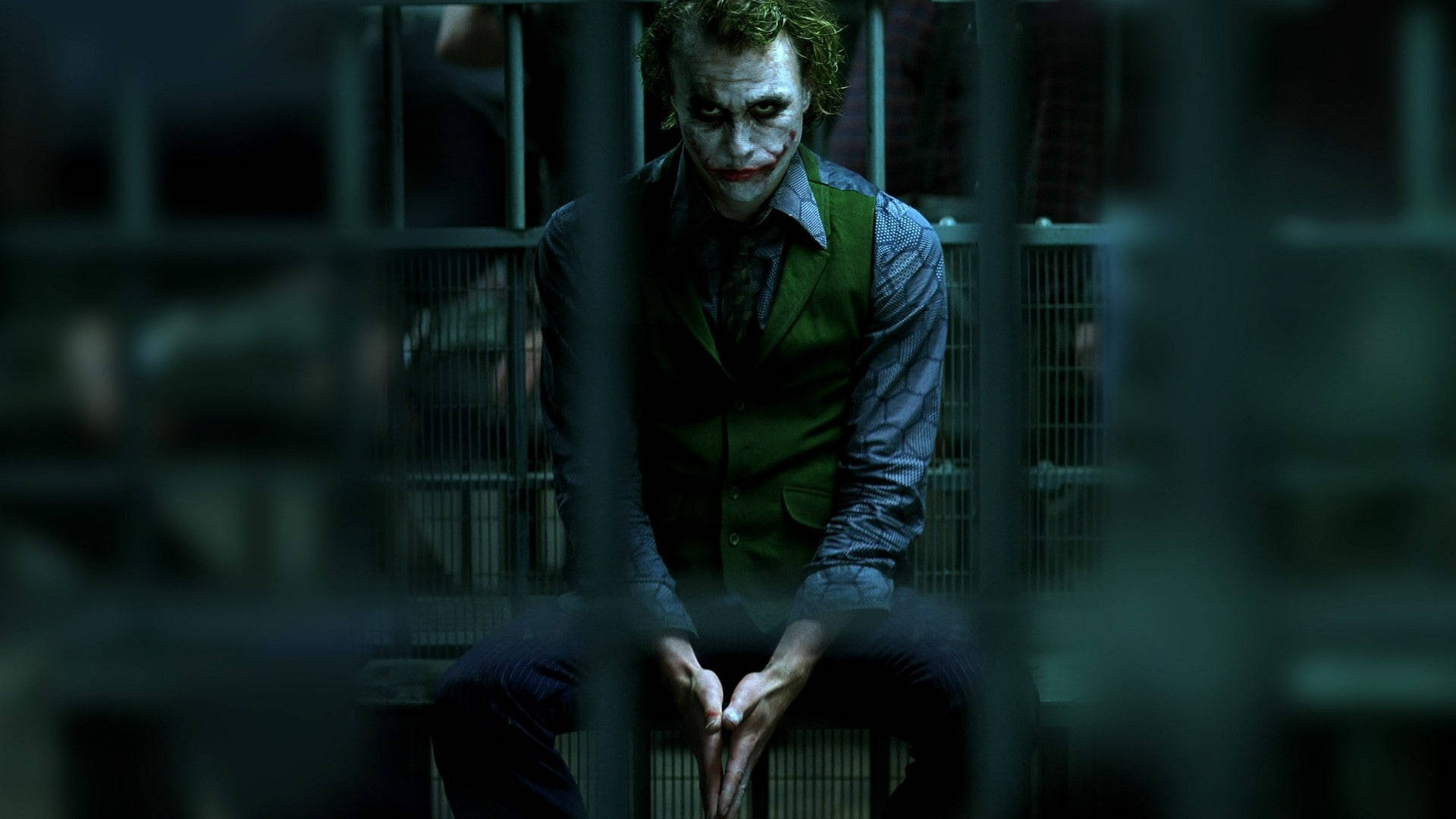 Heath Ledger Joker 桌布 壁紙 — 小丑坐在昏暗牢房的鐵欄後，身穿標誌性的綠色背心，臉上塗著白色顏料。