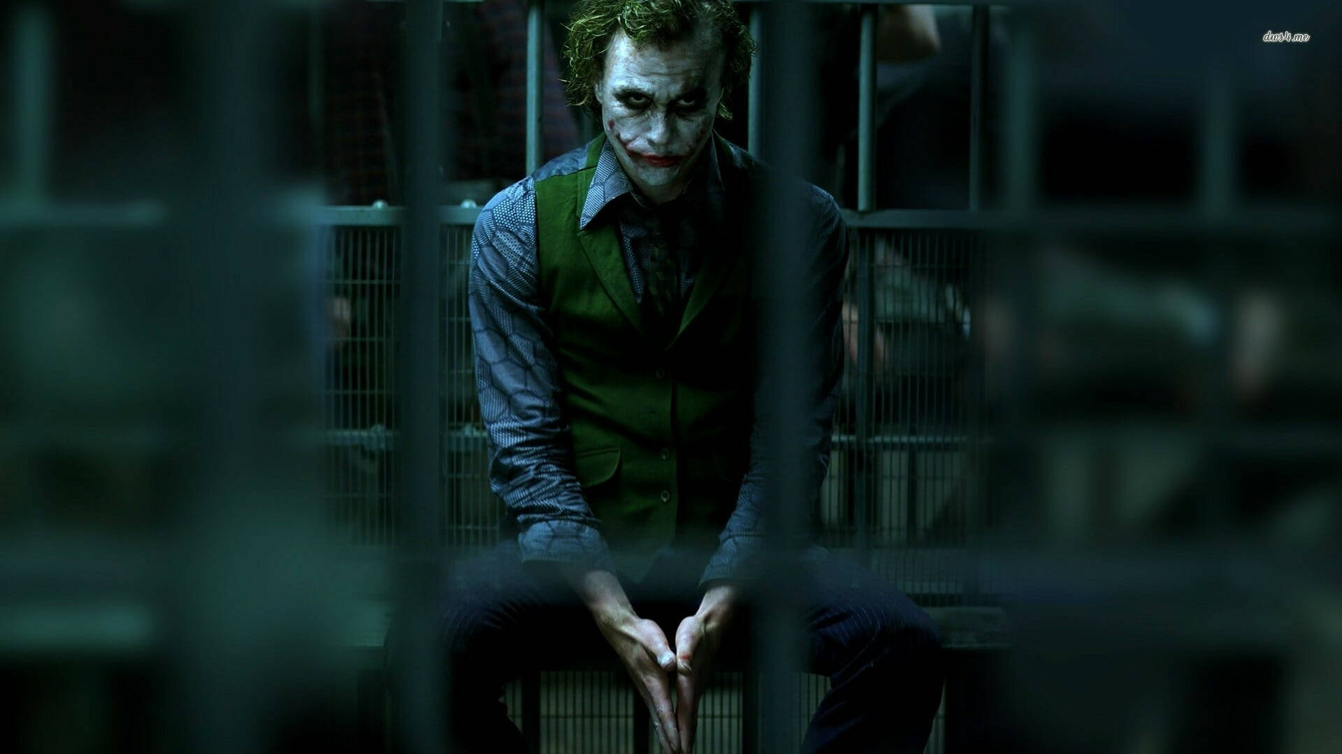 Why So Serious Joker 桌布 壁紙 — 小丑坐在昏暗牢房的鐵欄後，身穿標誌性的綠色背心，臉上塗著白色顏料。